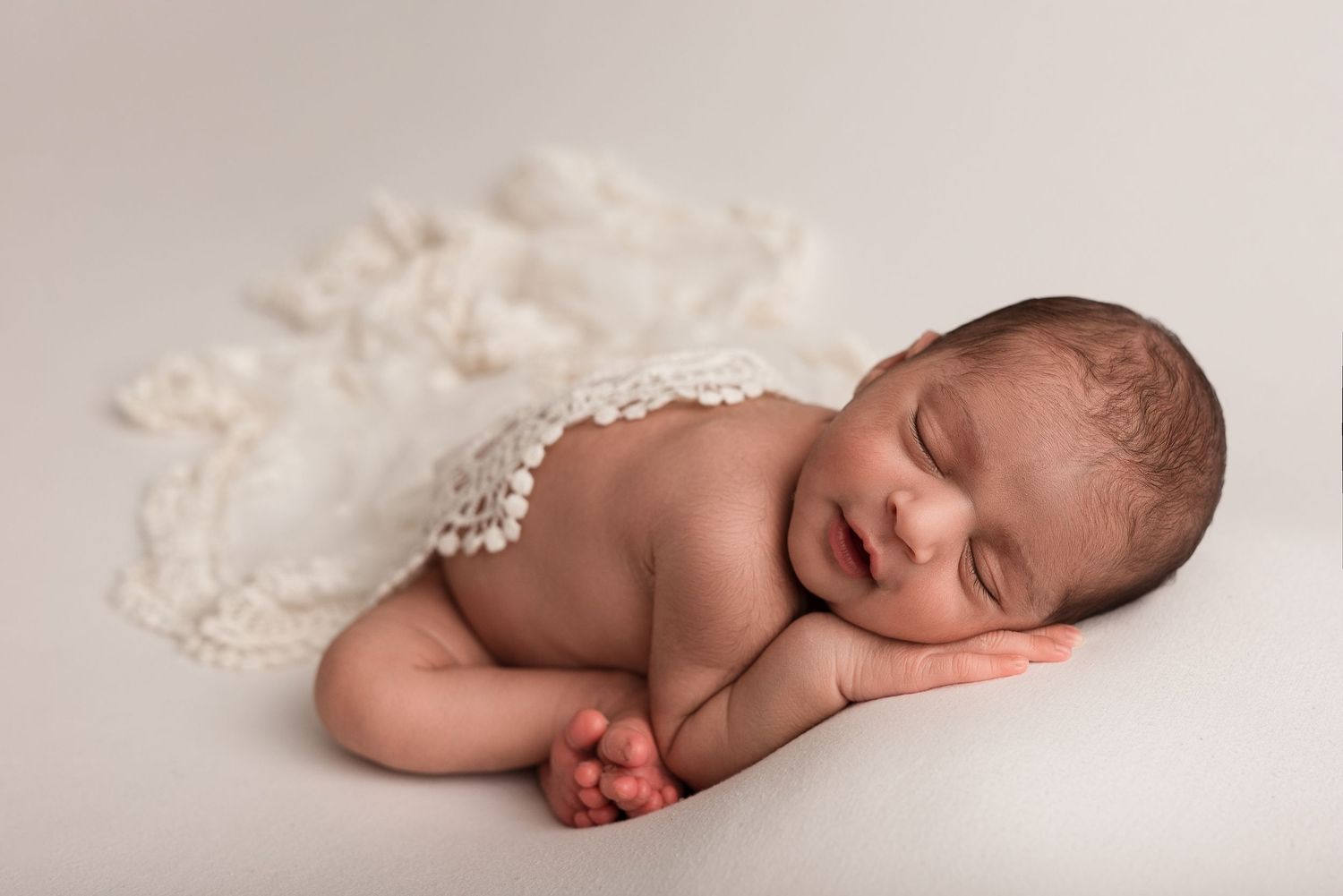 Peaceful newborn baby sleeping curled up on a white blanket with a delicate lace wrap, soft studio photography.