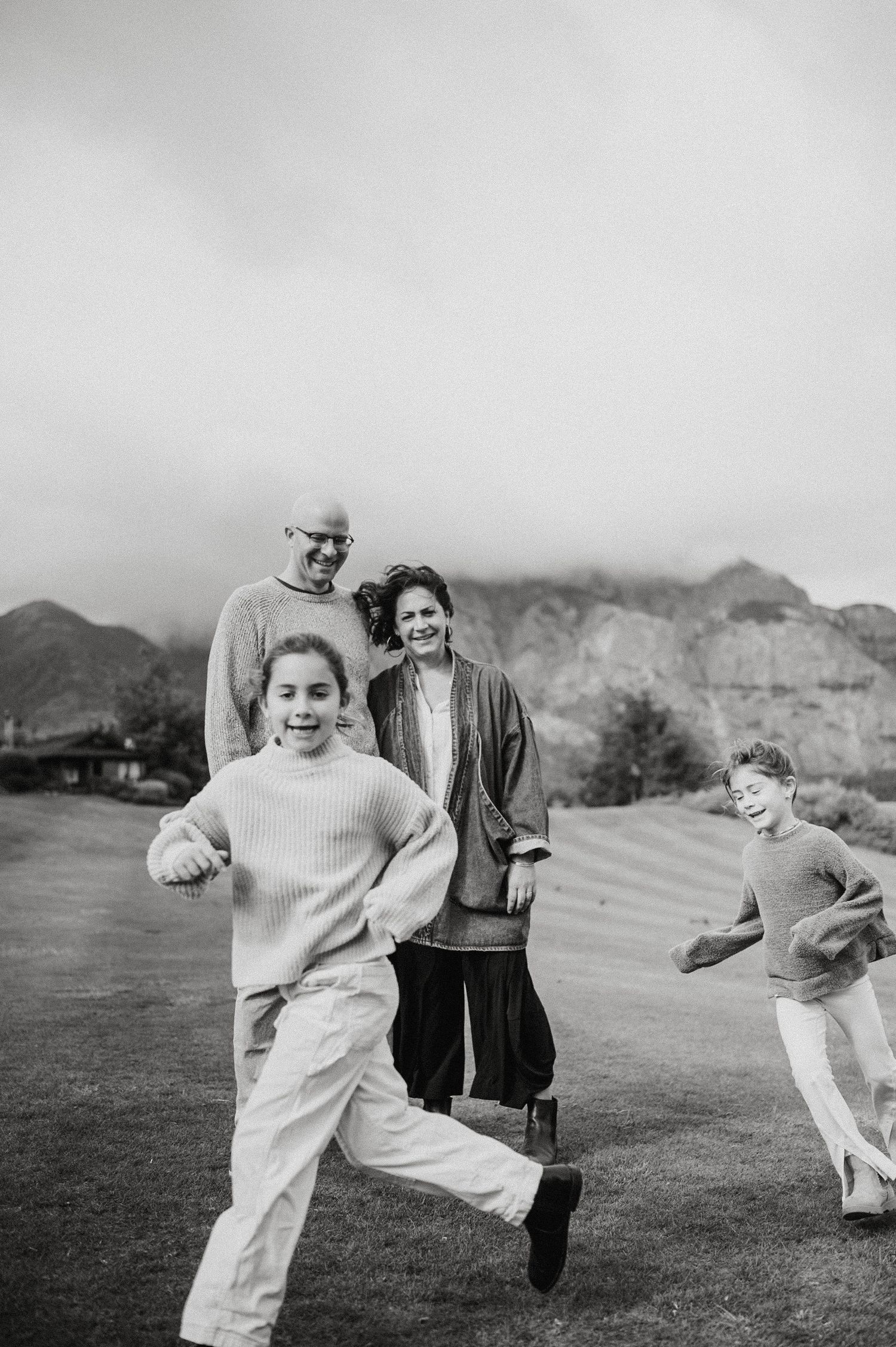 A black and white photo of people running and playing together on a grassy field with mountains in the background.