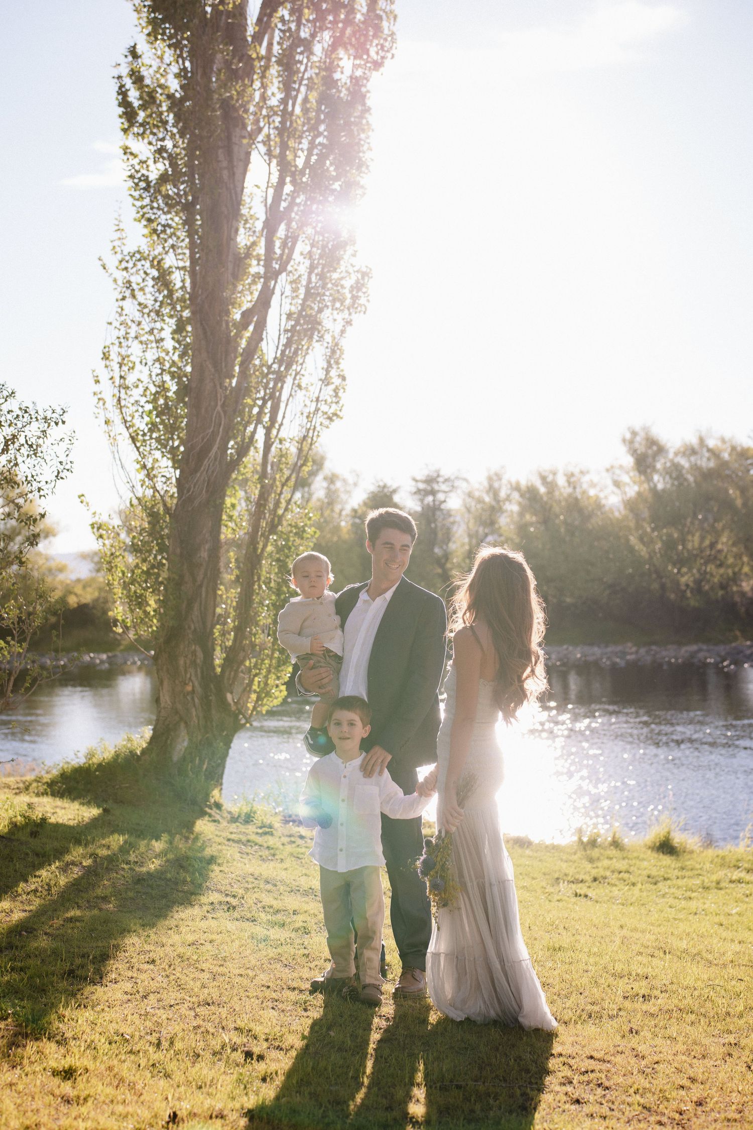 Family of four posing by a sunlit river, parents holding children, surrounded by lush green trees on a bright sunny day.
