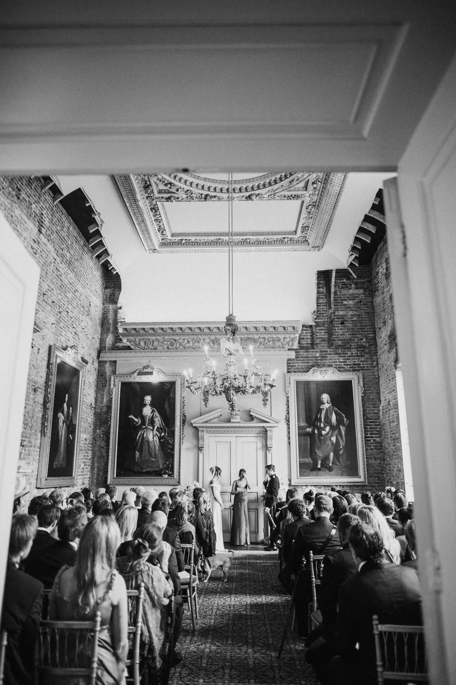 Black and white photo of an elegant wedding ceremony in a grand hall with ornate ceiling, chandelier, and large portrait paintings.
