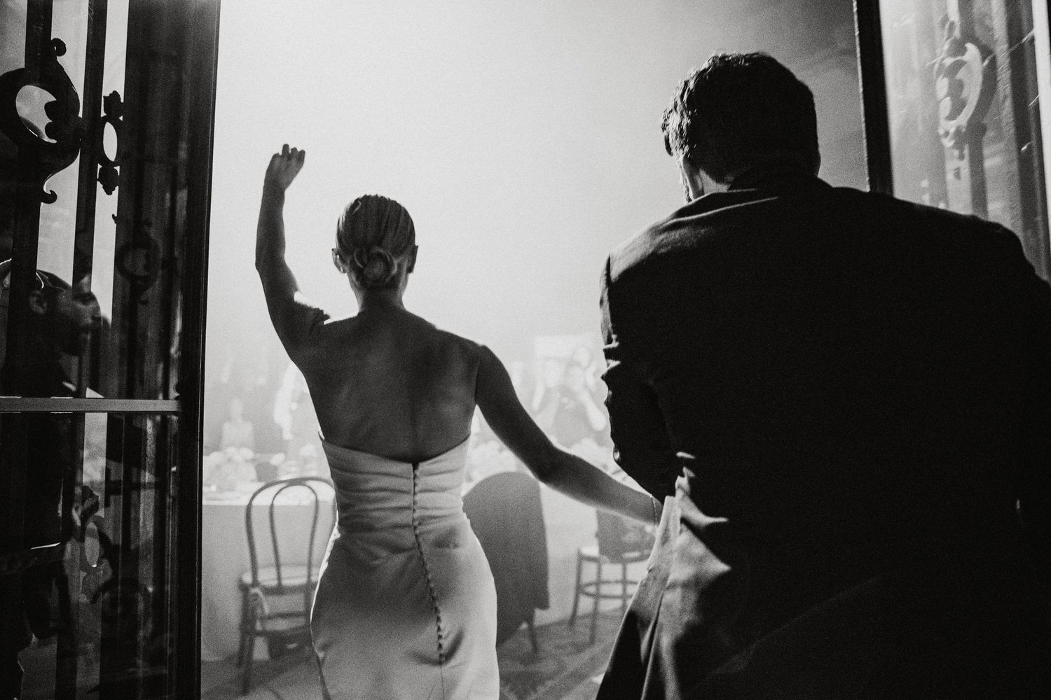 Black and white photo of bride in backless gown waving at wedding guests while groom watches from behind.