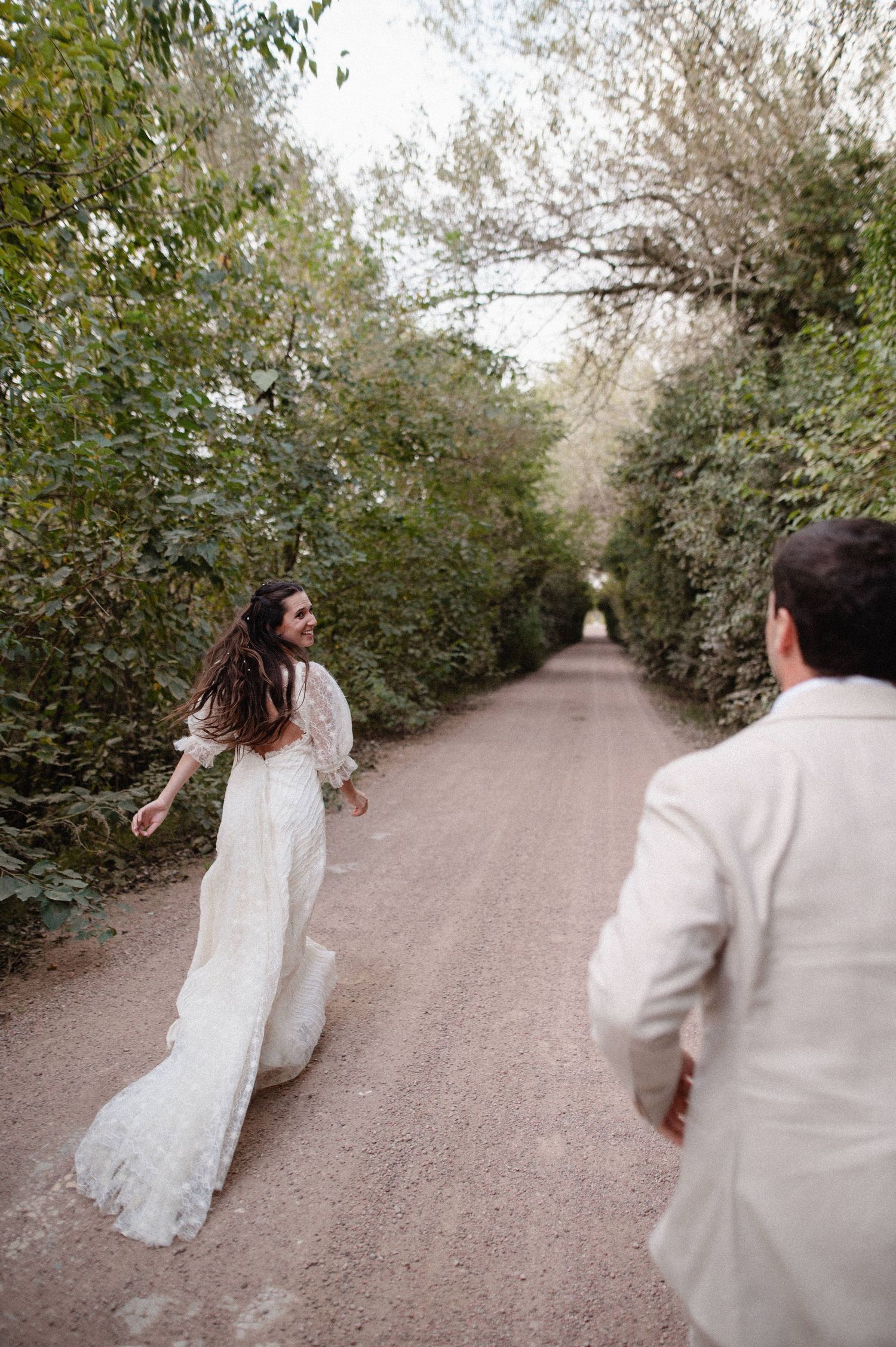 Bride in lace gown runs toward groom on a tree-lined dirt path during outdoor wedding photo session.