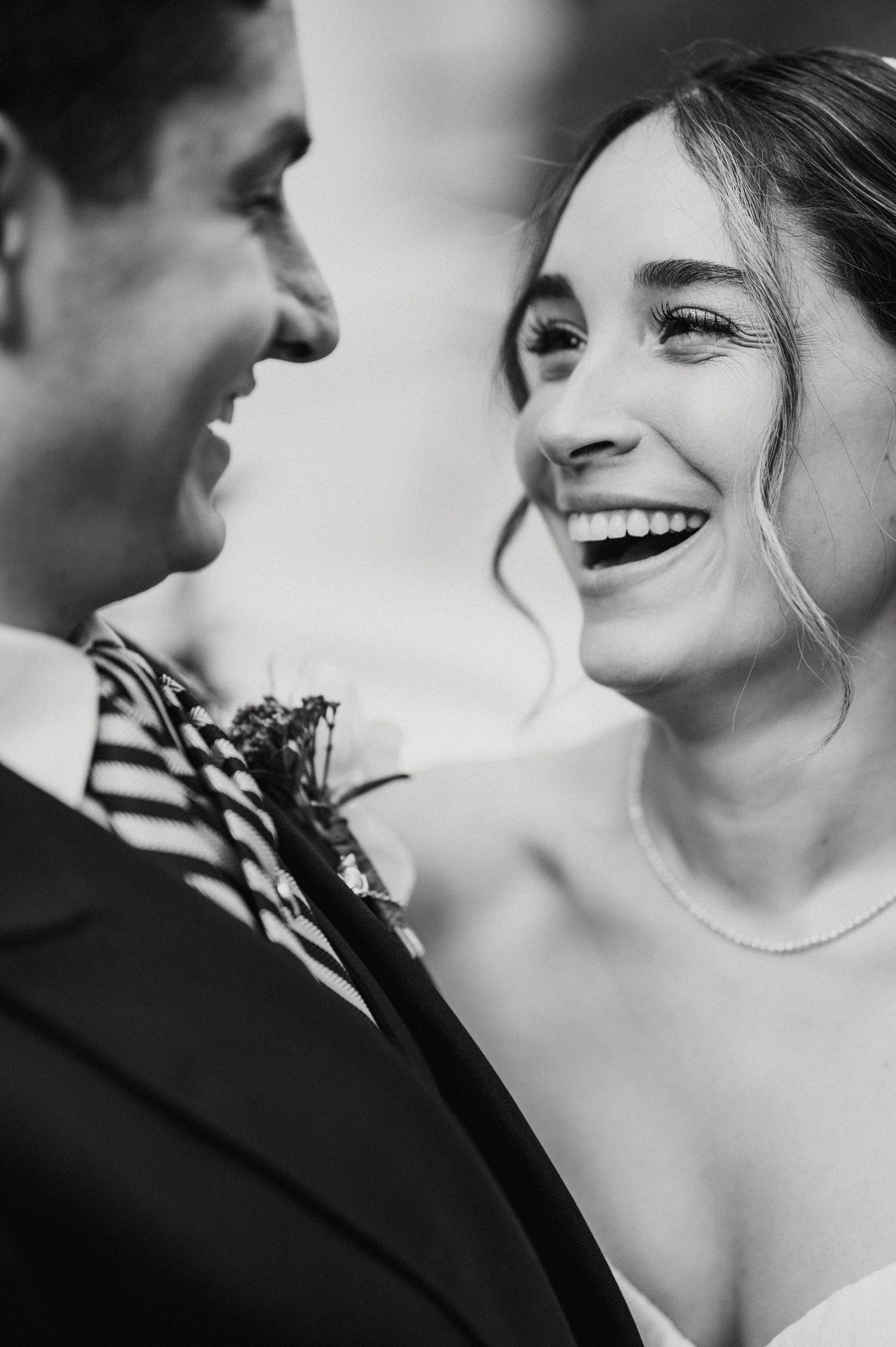 Black and white wedding photo of a laughing bride in pearls gazing at her groom in a striped tie with boutonniere.