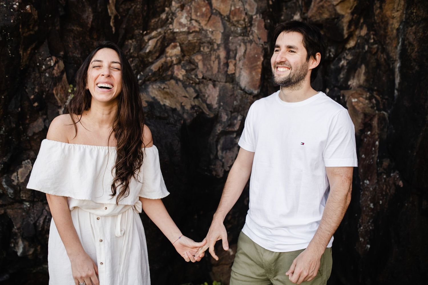 A couple in white outfits laughs together during a romantic photoshoot against a dark rocky background.
