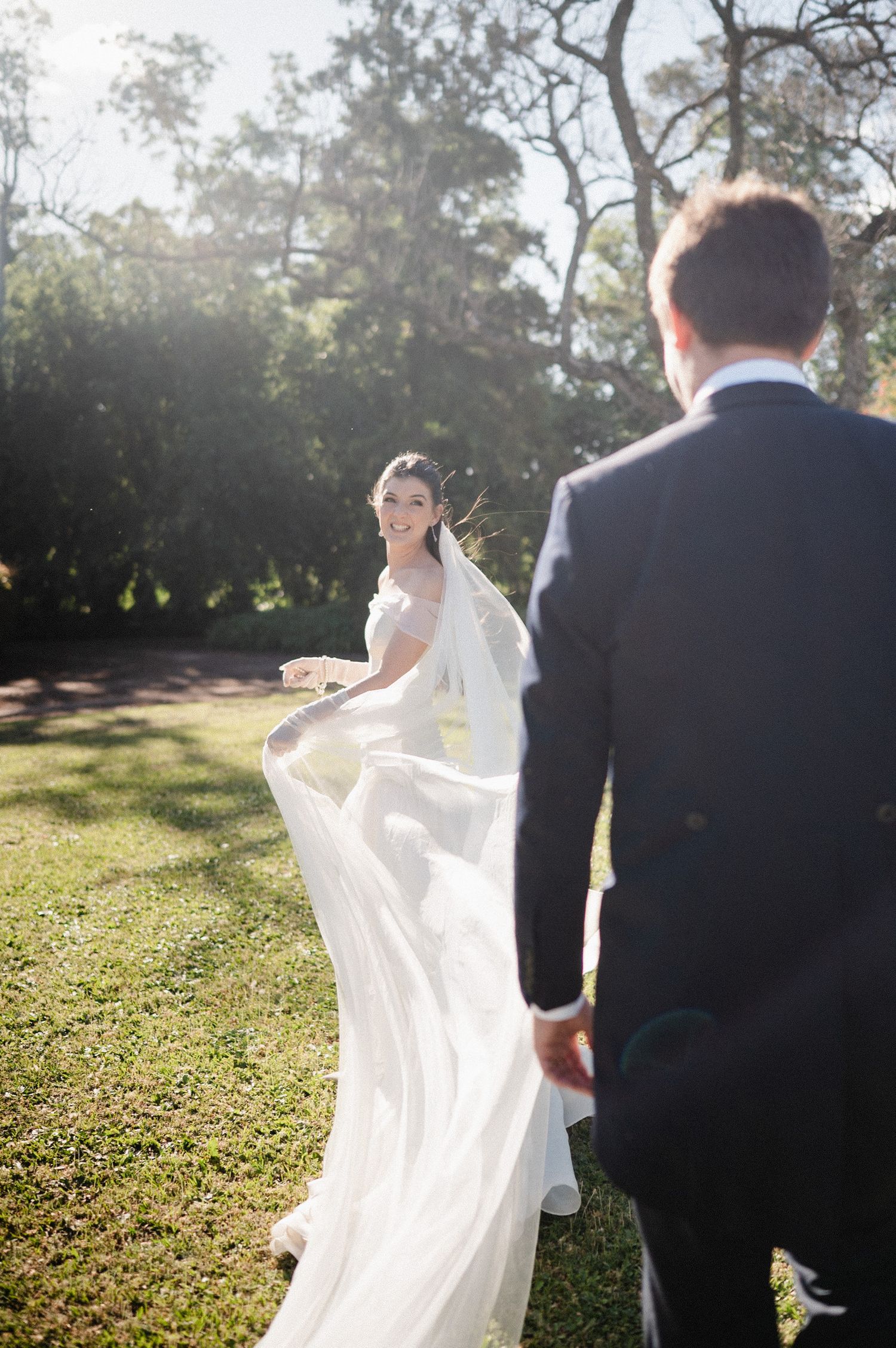 A bride and groom walk together in a sun-drenched garden as her white dress flows elegantly in the breeze.