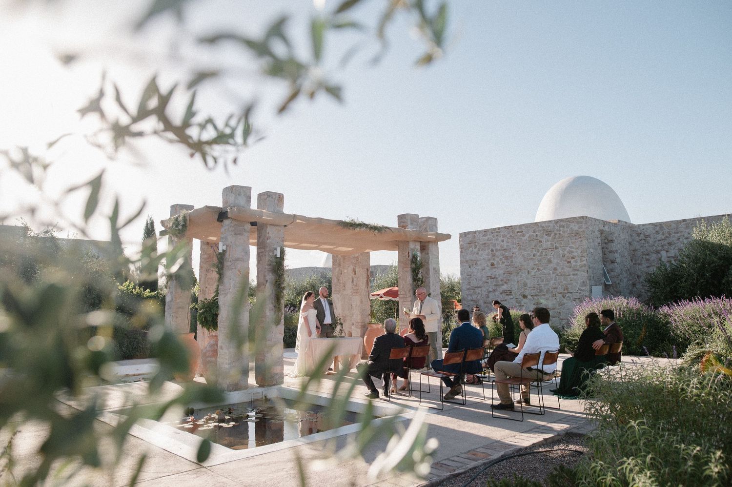Outdoor wedding ceremony at a stone venue with guests seated, olive trees, and a dome structure under clear blue sky.