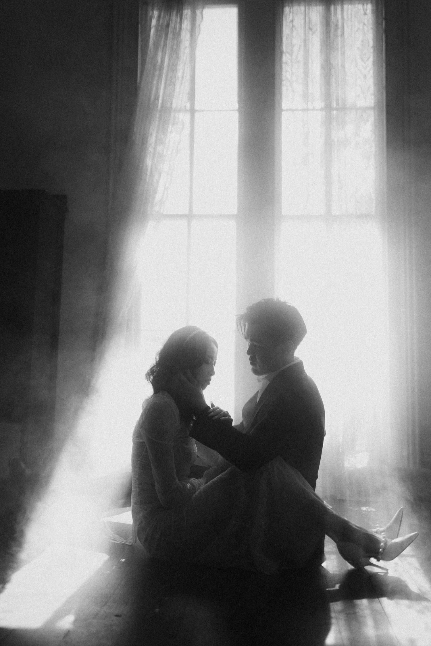Two silhouettes sit intimately by a bright window in black and white.