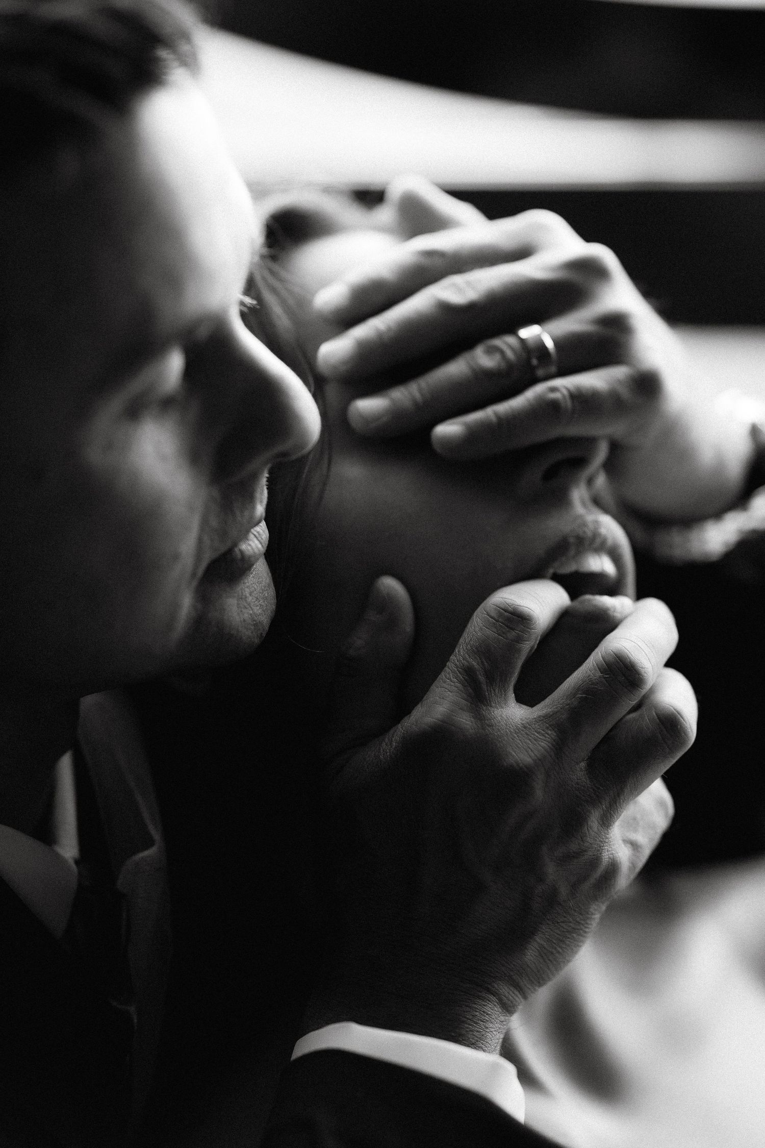 Black and white photo of a couple, man gently holding woman's face with both hands, wearing wedding rings.