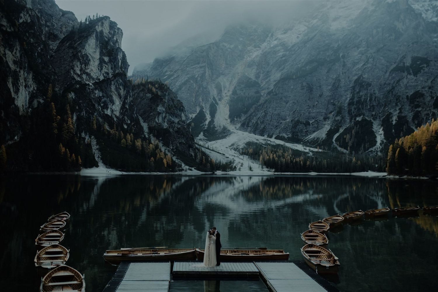 Couple standing on dock overlooking misty Lago di Braies lake with wooden boats and dramatic Dolomite mountains in Italy.