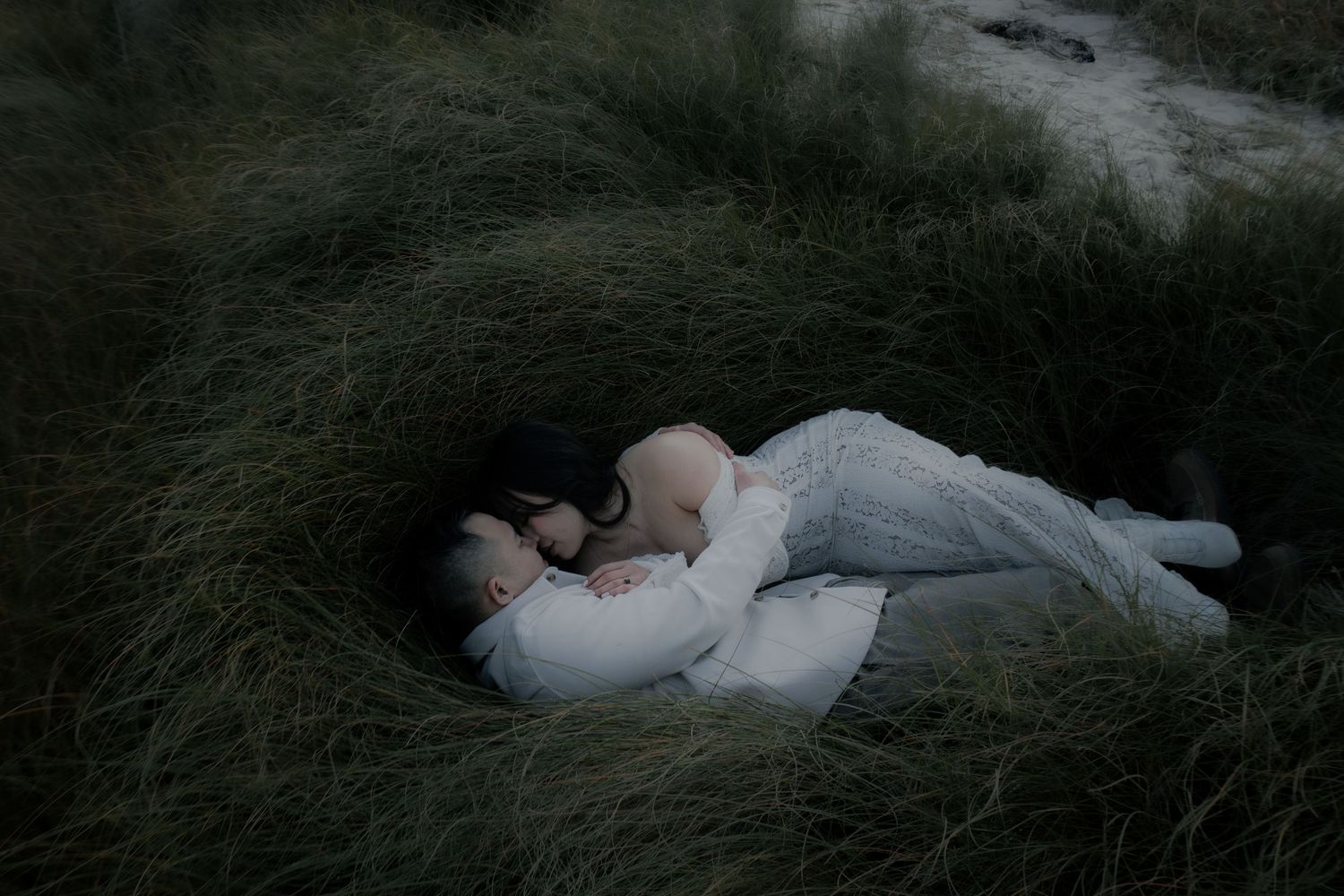 A couple in light colored clothing lies together intimately in tall grass at dusk.