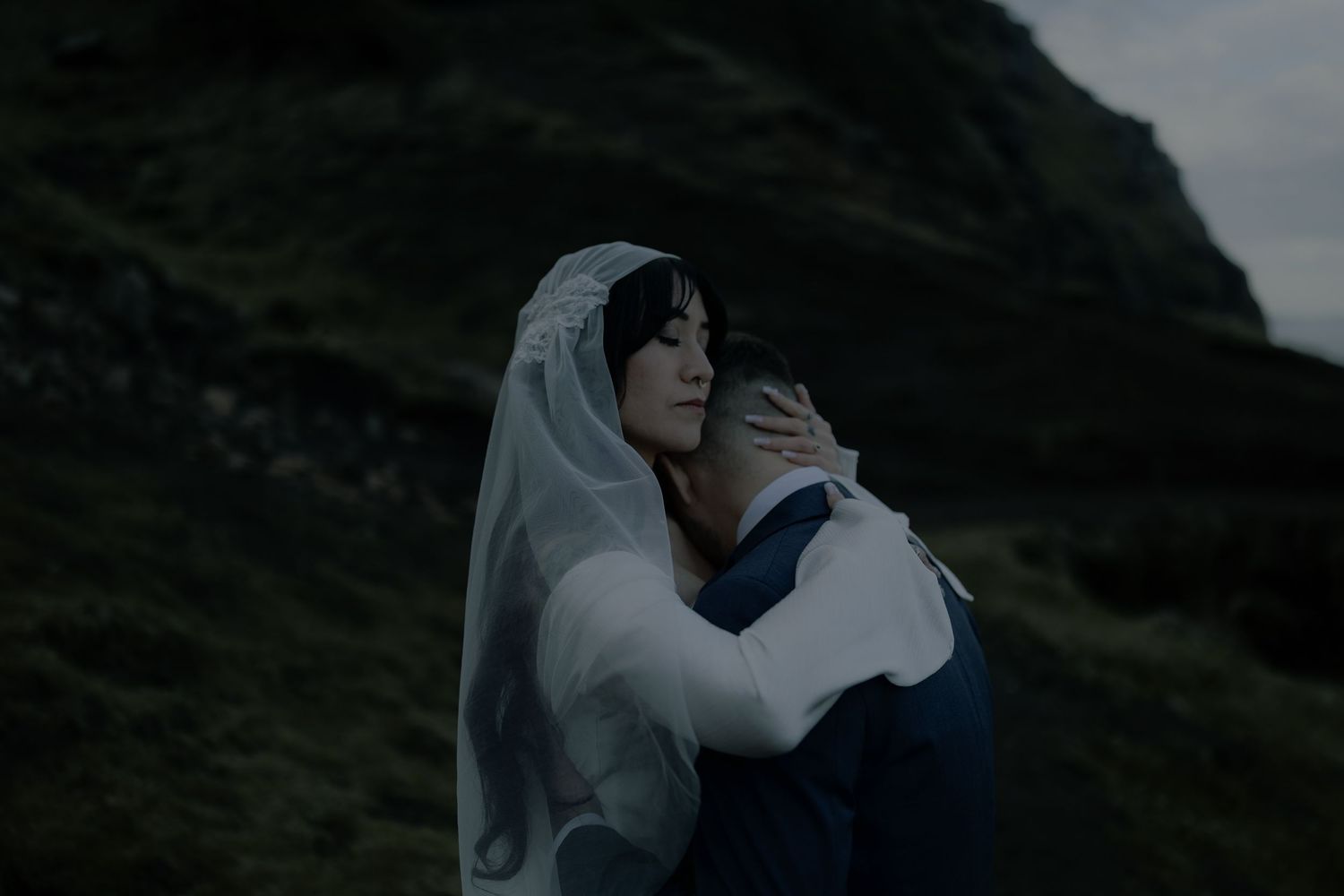 A bride in a white veil embraces a groom on a moody hillside in a romantic outdoor wedding portrait.