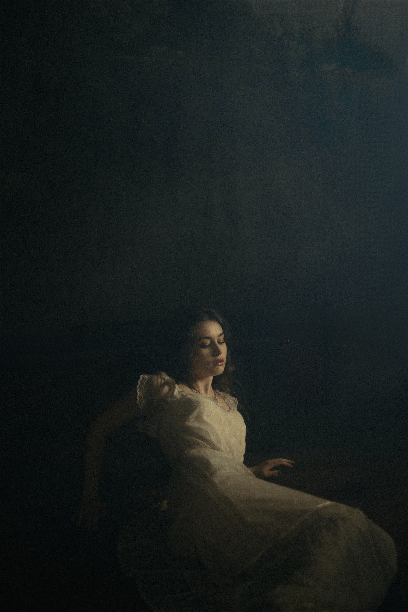 A dramatic moody portrait shows a figure in white reclining against a dark background in cinematic lighting.