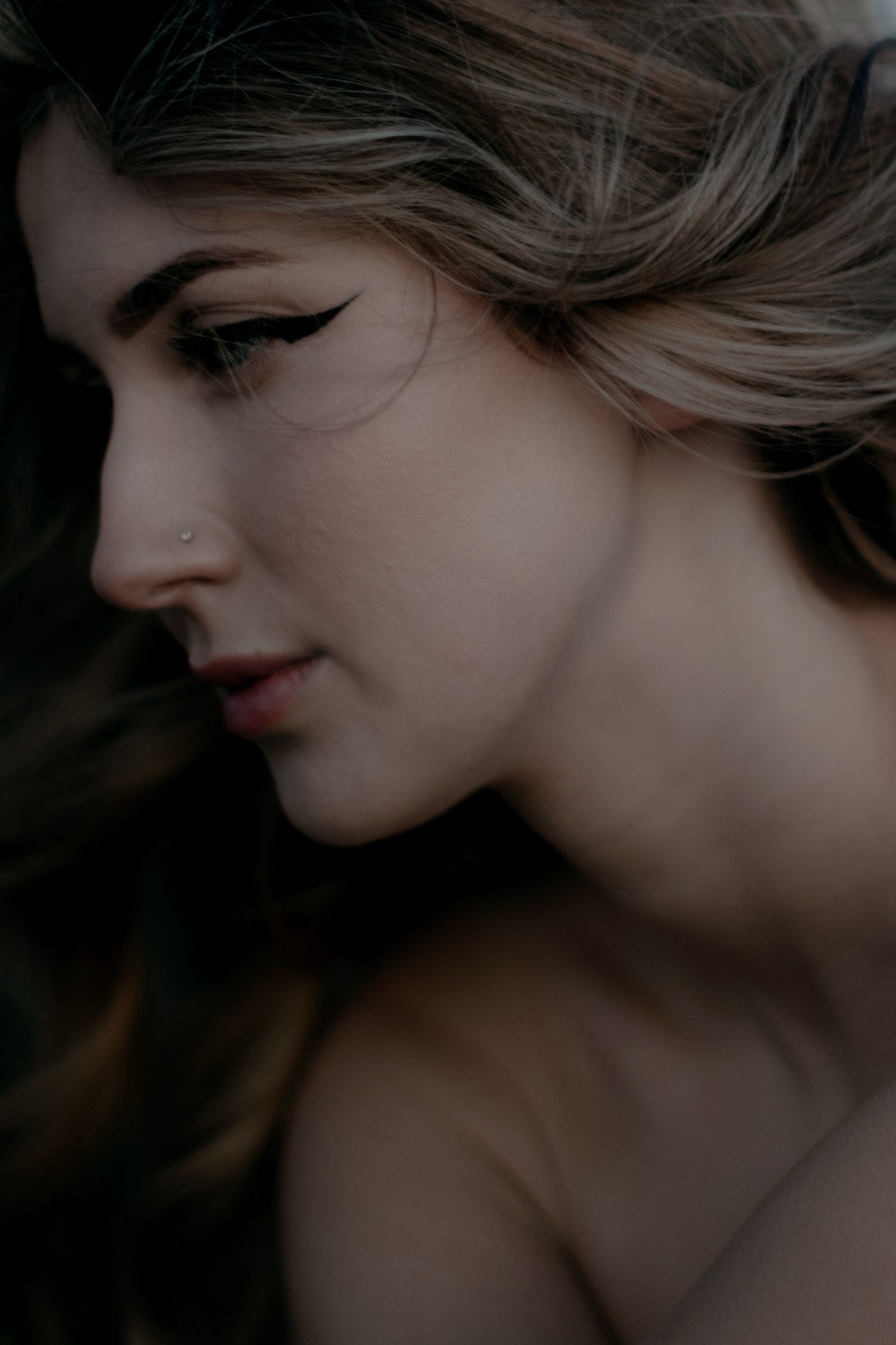 Close-up profile of a woman with winged eyeliner, nose ring, and wavy hair in a moody, dark artistic portrait.