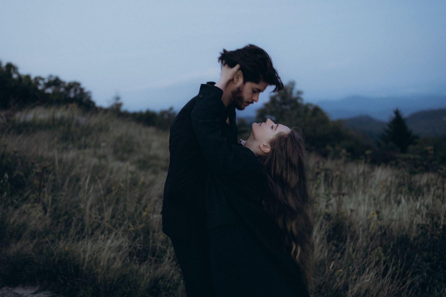 Romantic couple sharing an intimate moment at dusk in a scenic mountain meadow with misty hills in the background.