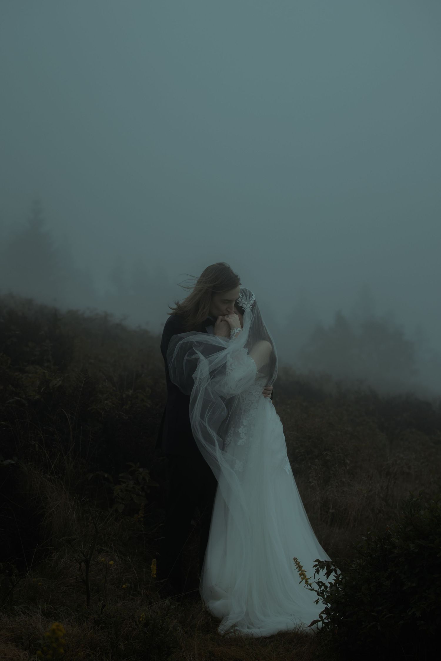 A bride and groom embrace in a misty foggy landscape, her white gown and veil flowing in the moody outdoor setting.