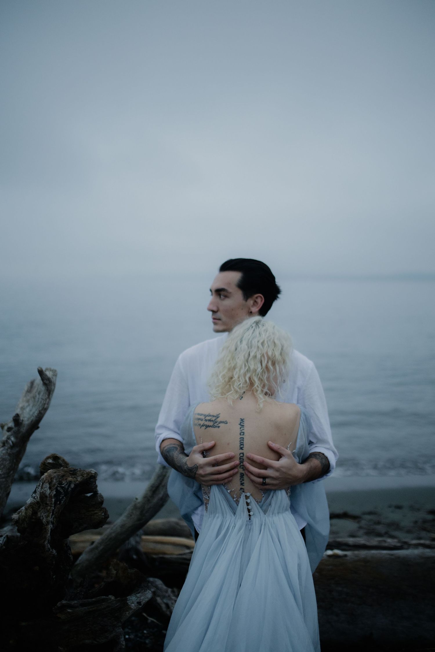 A couple embraces on a misty beach, with the person in white wearing a flowing dress against a moody gray backdrop.