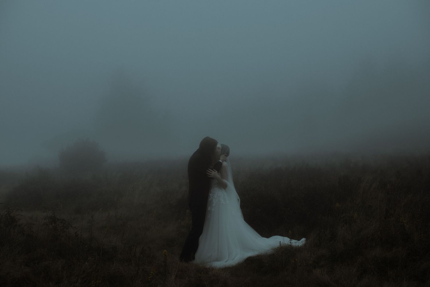 A couple shares a romantic embrace in a misty foggy field, bride in white gown and groom in dark suit.