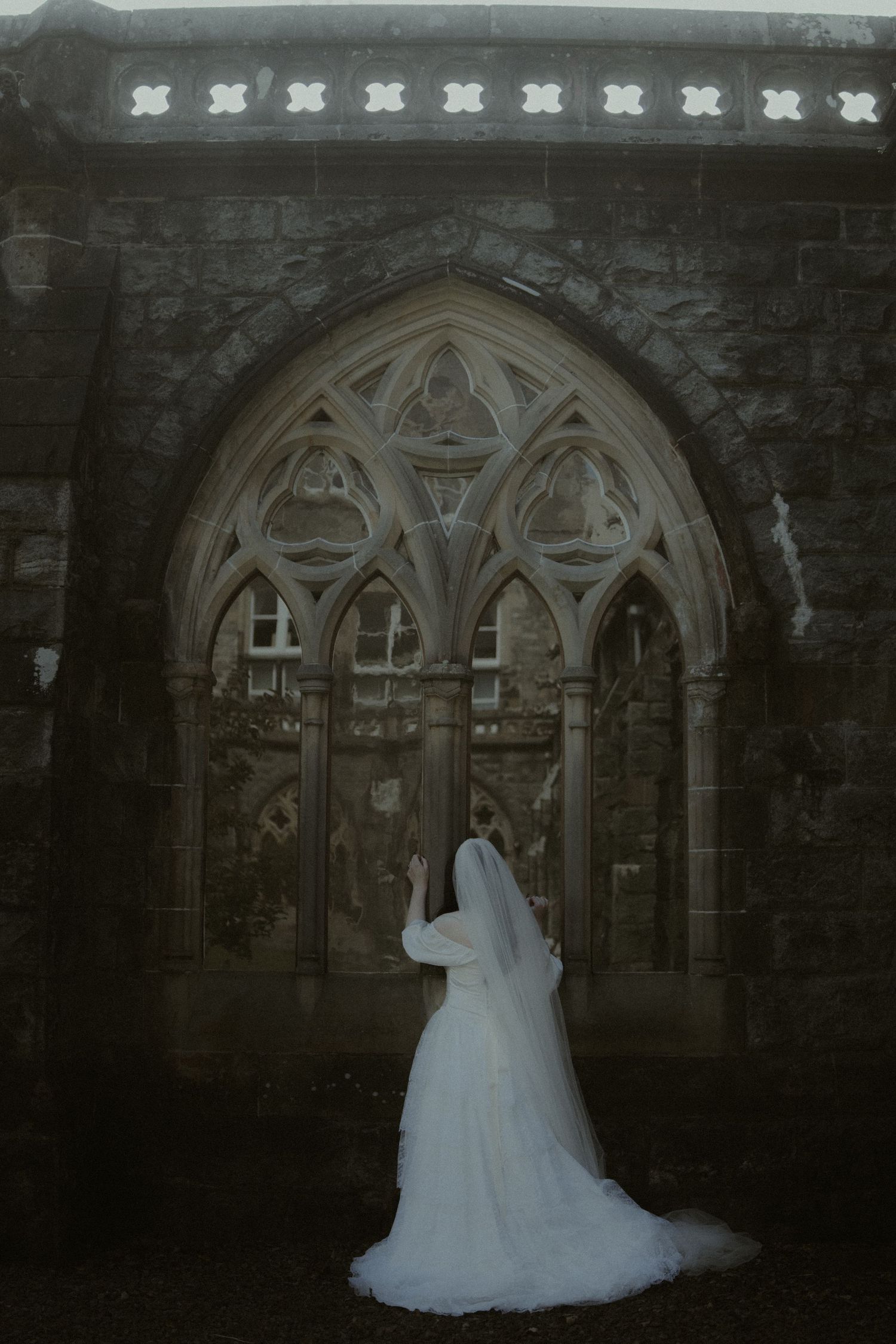 Bride in white gown and veil stands before ornate Gothic stone church doorway in moody, atmospheric setting.