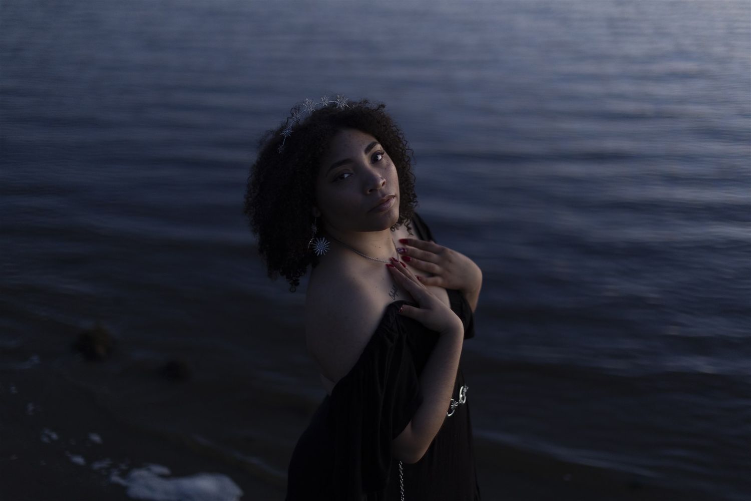 A woman in a black off-shoulder dress poses gracefully by the water at dusk, hands crossed over her chest.