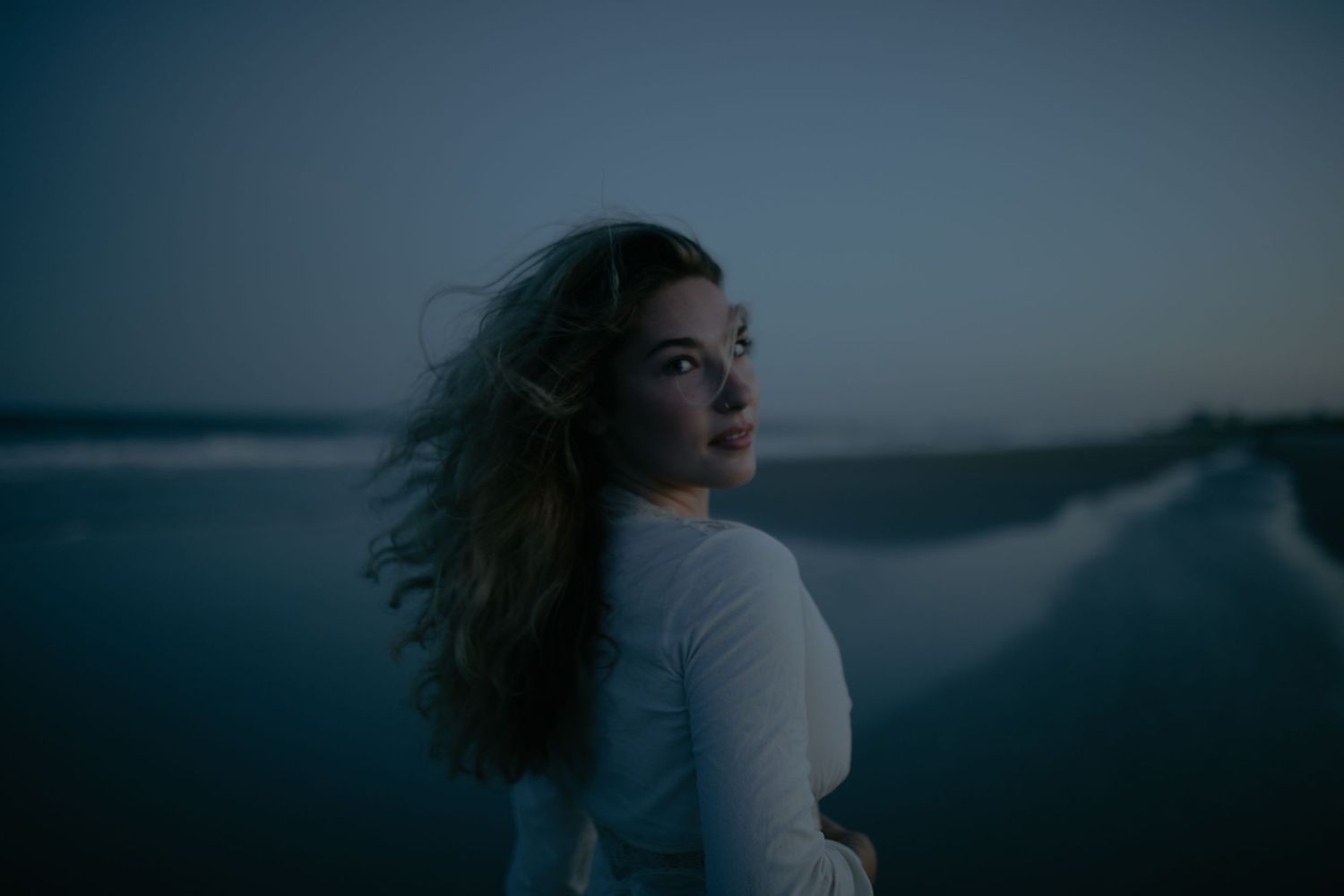 Young woman with curly hair in white top gazes upward at dusk on a serene beach with soft blue twilight sky.