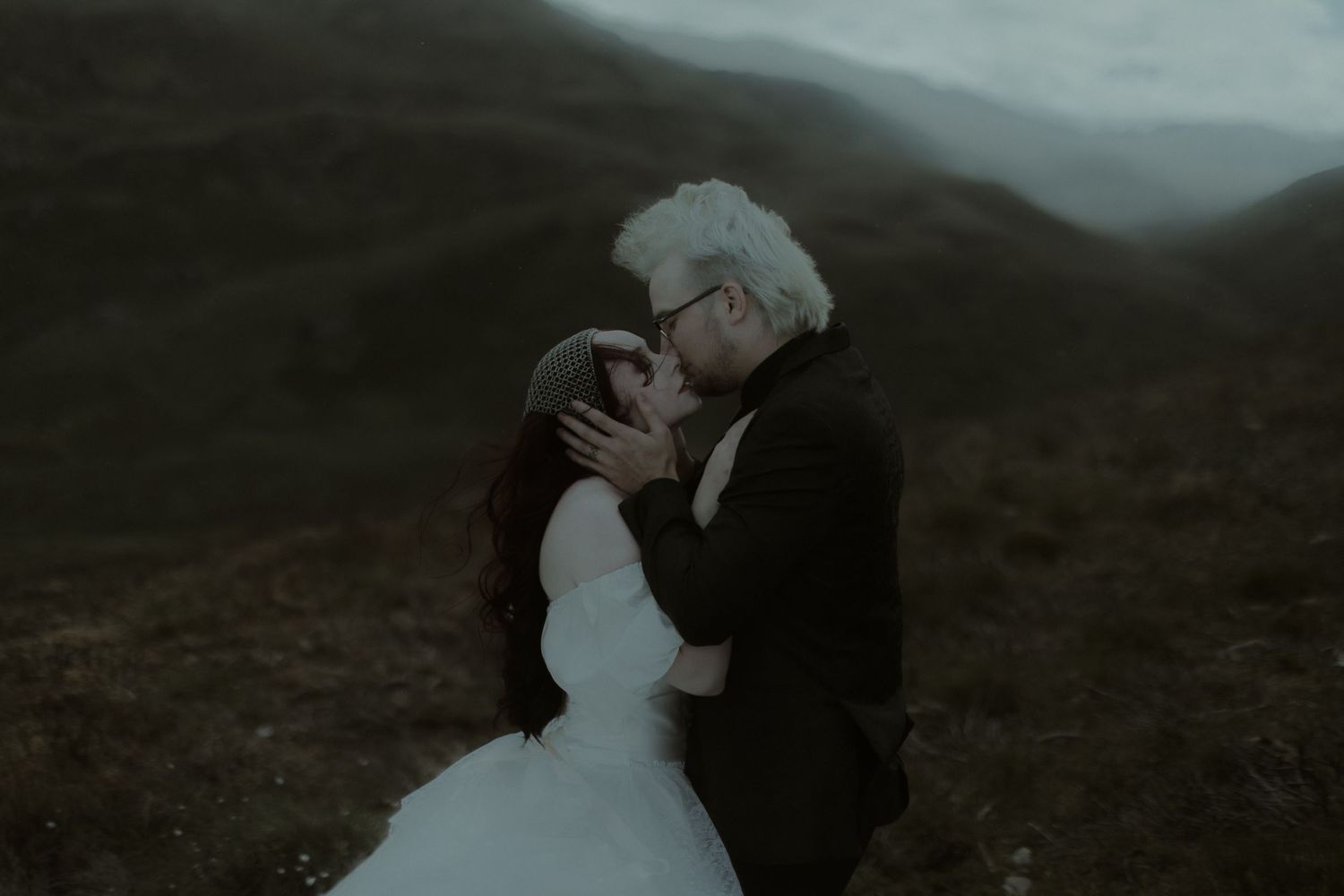 A couple shares a romantic kiss outdoors in a misty mountain landscape, bride in white gown and groom with silver hair.
