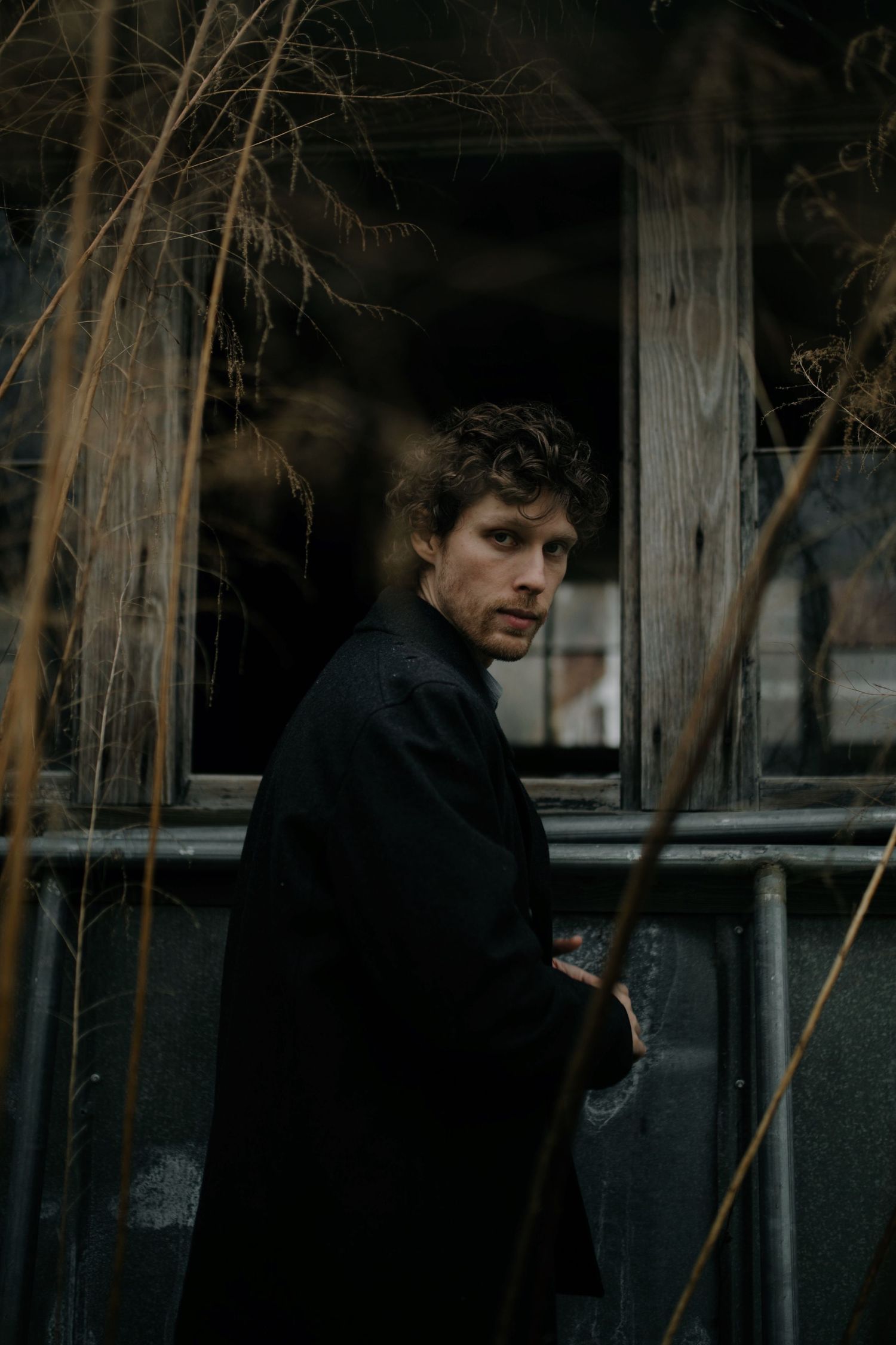 A brooding man in a black coat stands near a weathered wooden window surrounded by dry branches.