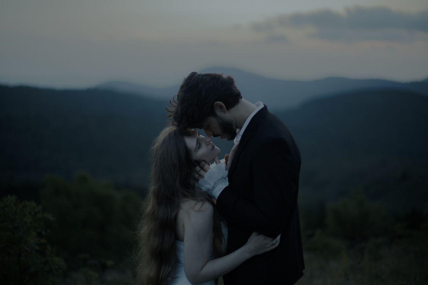 Romantic couple embracing at dusk with misty mountain forest backdrop in soft twilight light.