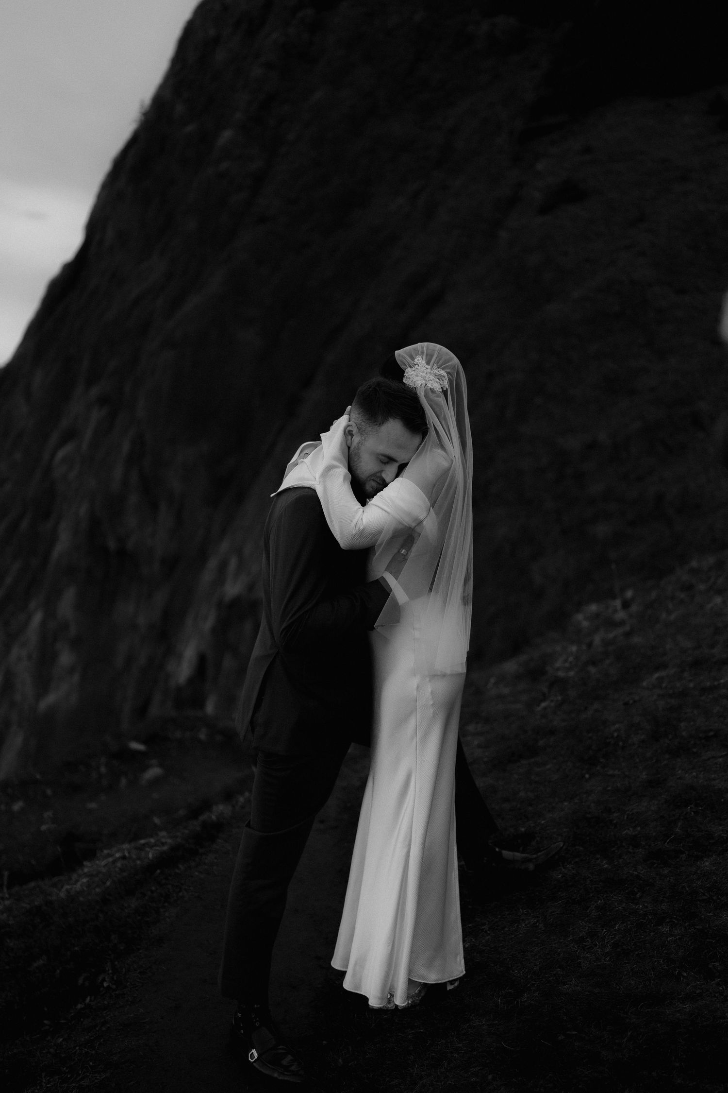 Photo of a bride and groom embracing near a rocky cliff, her veil flowing gracefully in the wind during their Oregon Coast elopement.