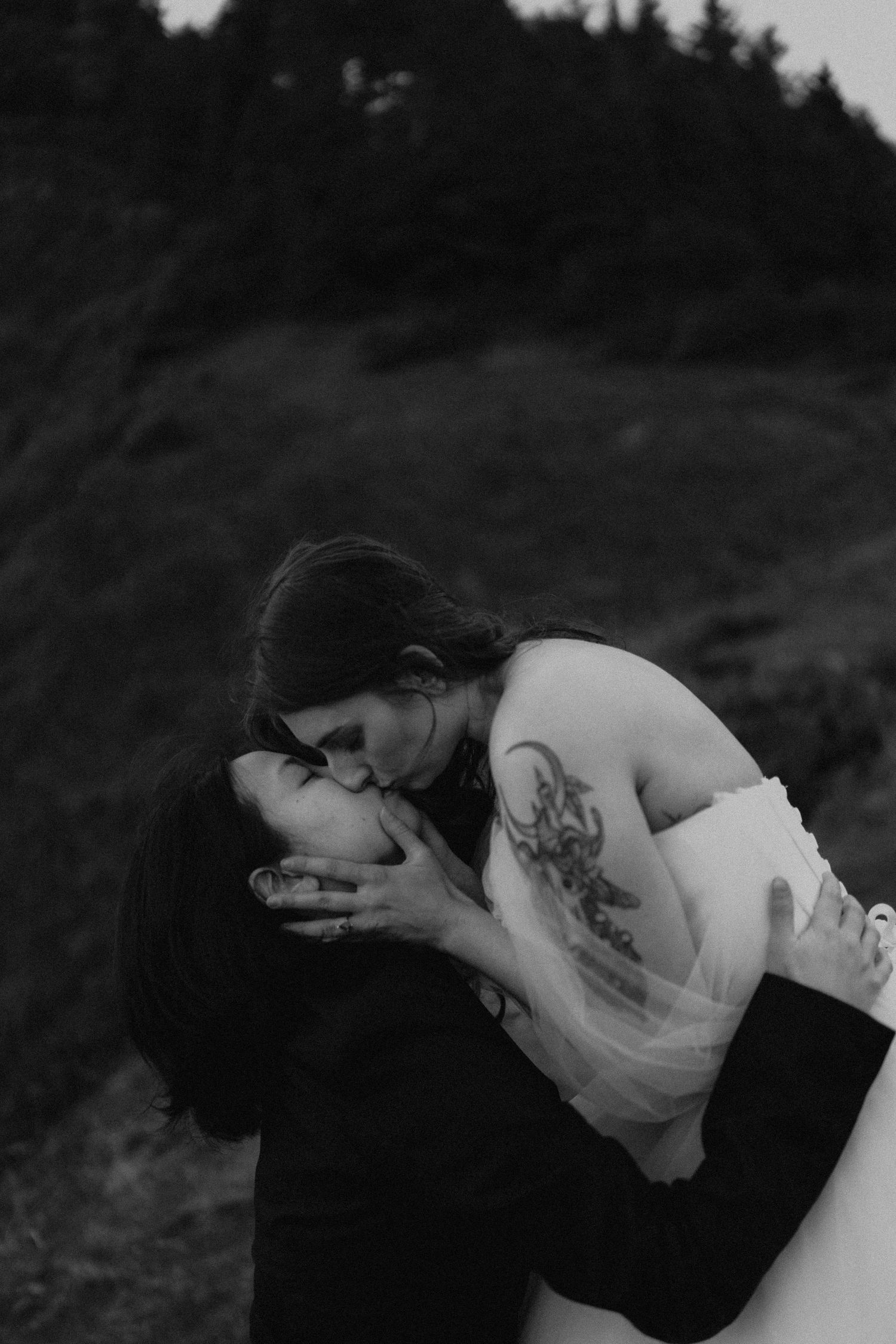 Black and white photo of a couple sharing a romantic dip kiss outdoors, bride in white dress with tattoo visible.