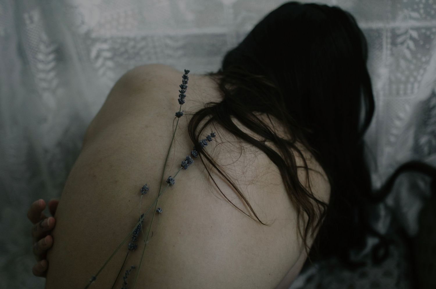 Person with dark hair showing a delicate floral spine tattoo on their bare back against a soft curtain backdrop.