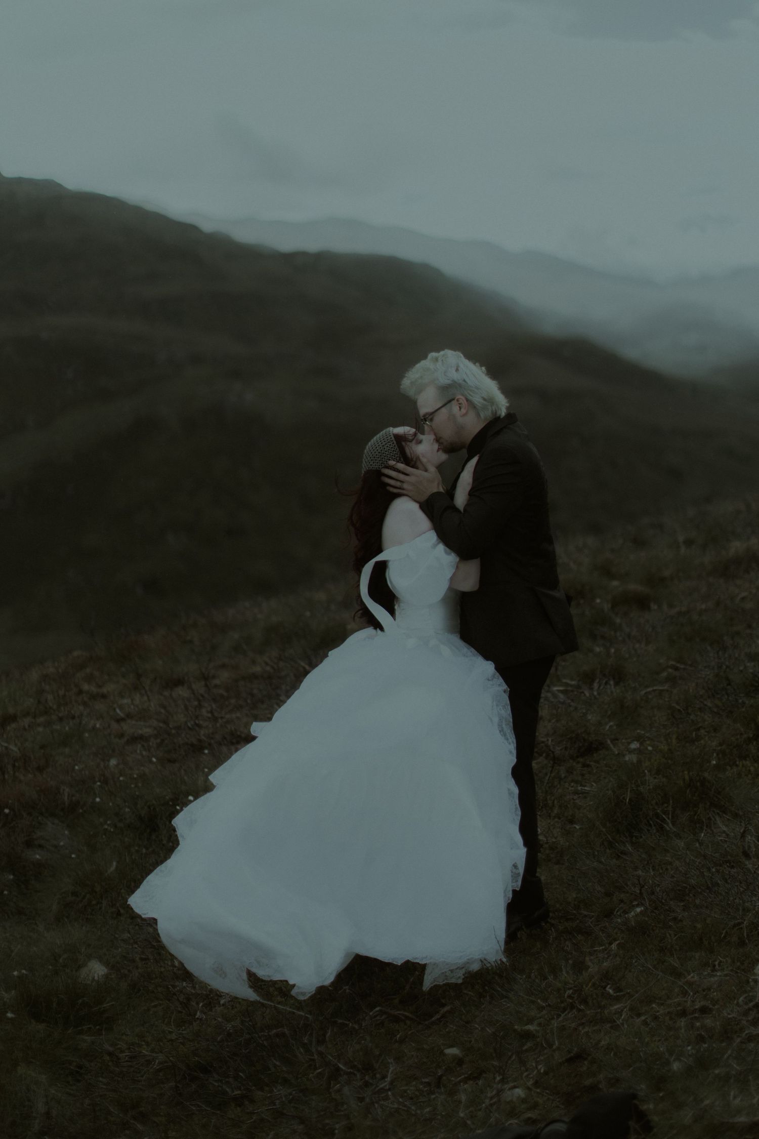 A couple shares a romantic kiss in wedding attire on a misty mountain hillside, bride in white gown, groom in dark suit.