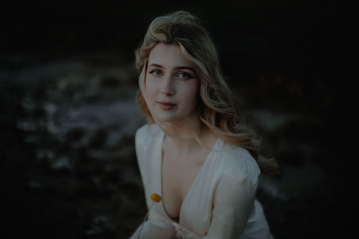 Young woman with blonde hair in a white dress poses outdoors near a rocky stream in a moody portrait.