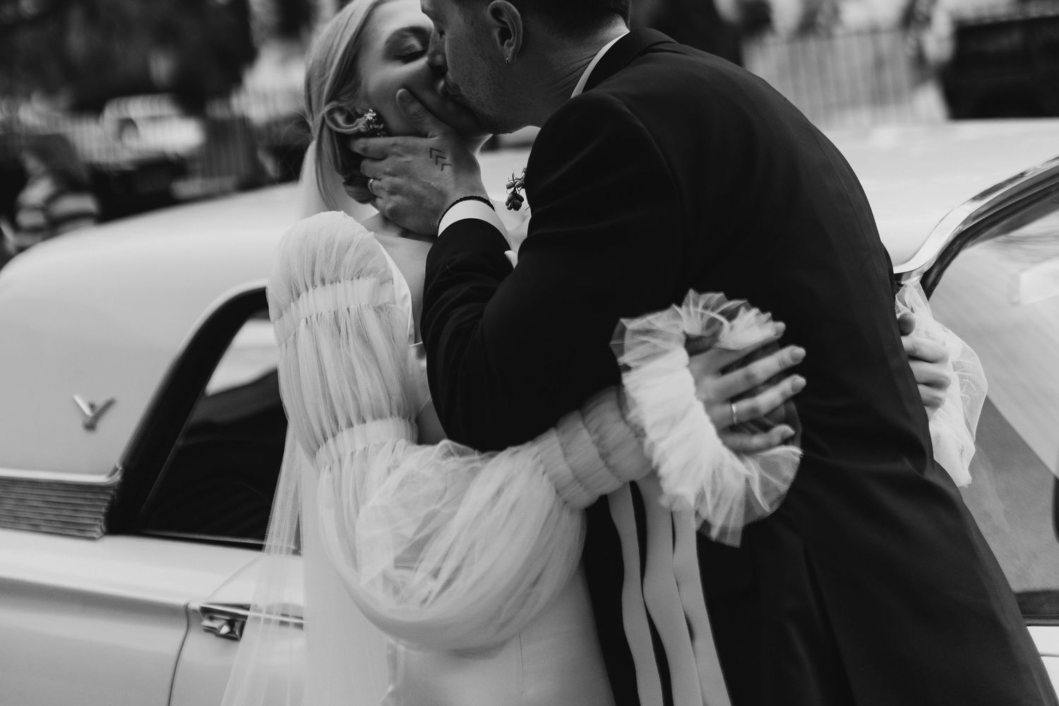 Black and white photo of bride and groom sharing a kiss beside a vintage white car on their wedding day.