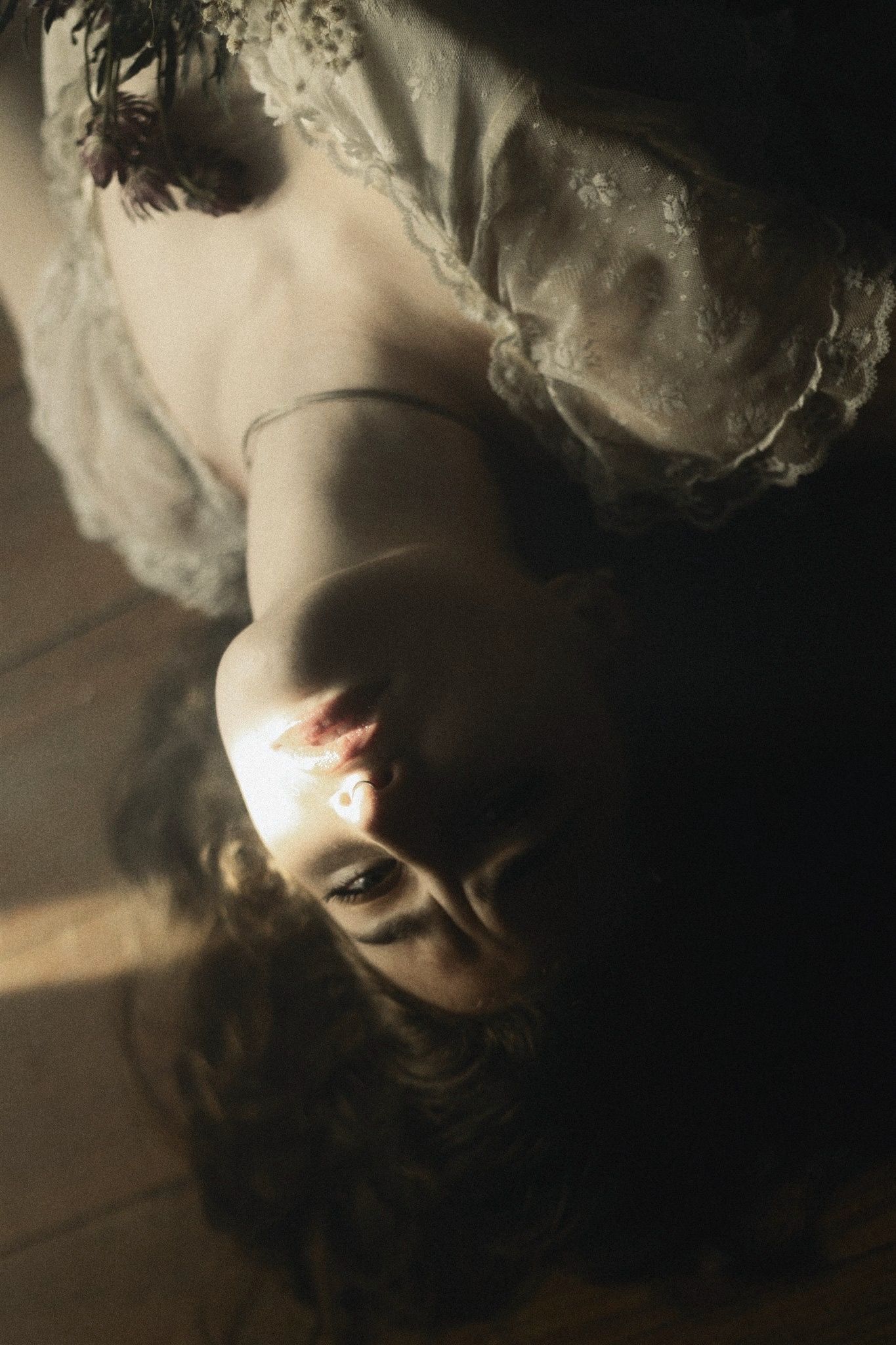 Artistic upside-down portrait of a woman in lace dress with flowers, dramatic moody lighting on wooden floor.