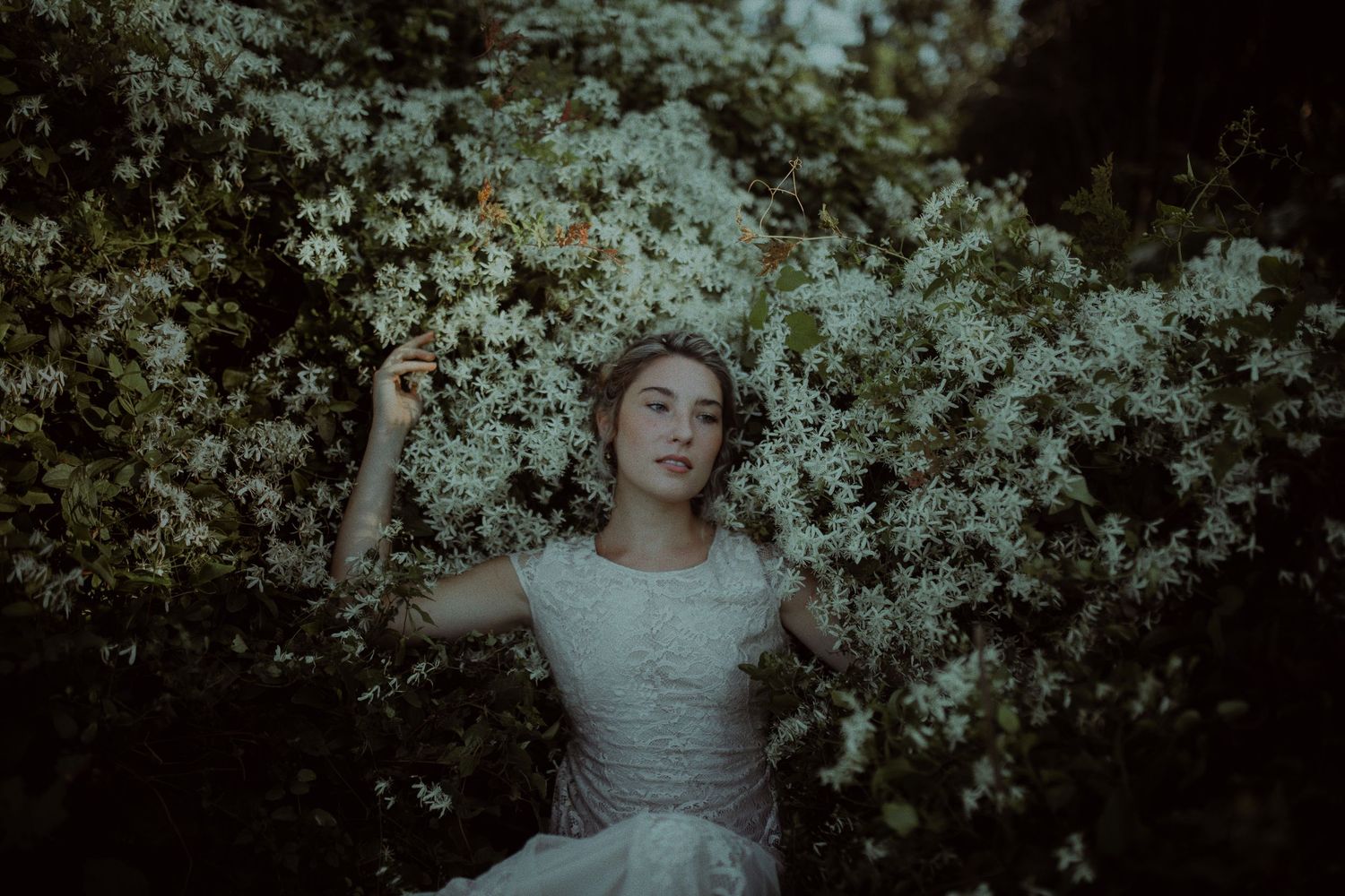 Young woman in white dress surrounded by blooming white flowers in a lush green garden setting.