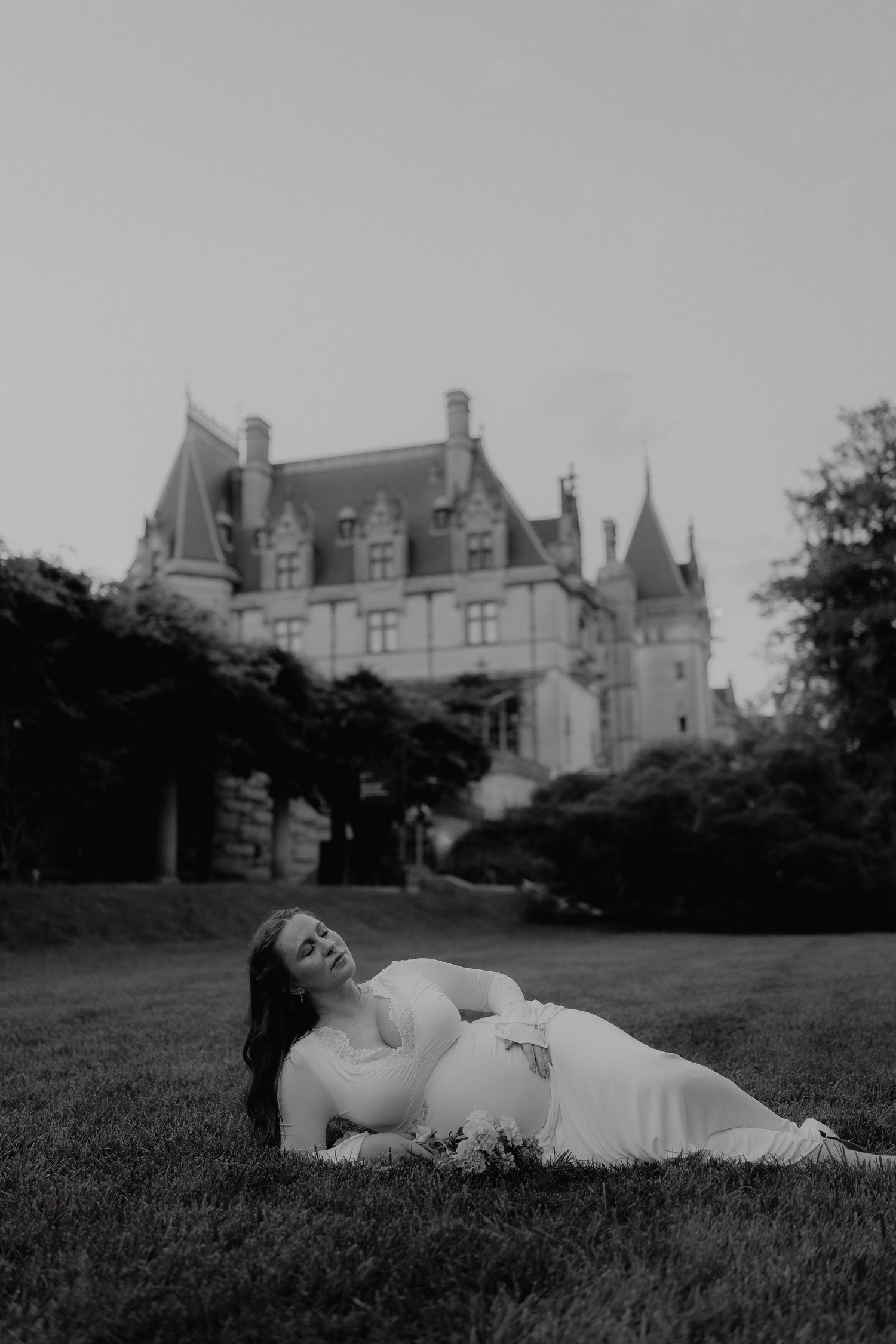Pregnant woman in white dress lying on grass before a grand castle in a black and white maternity photo.