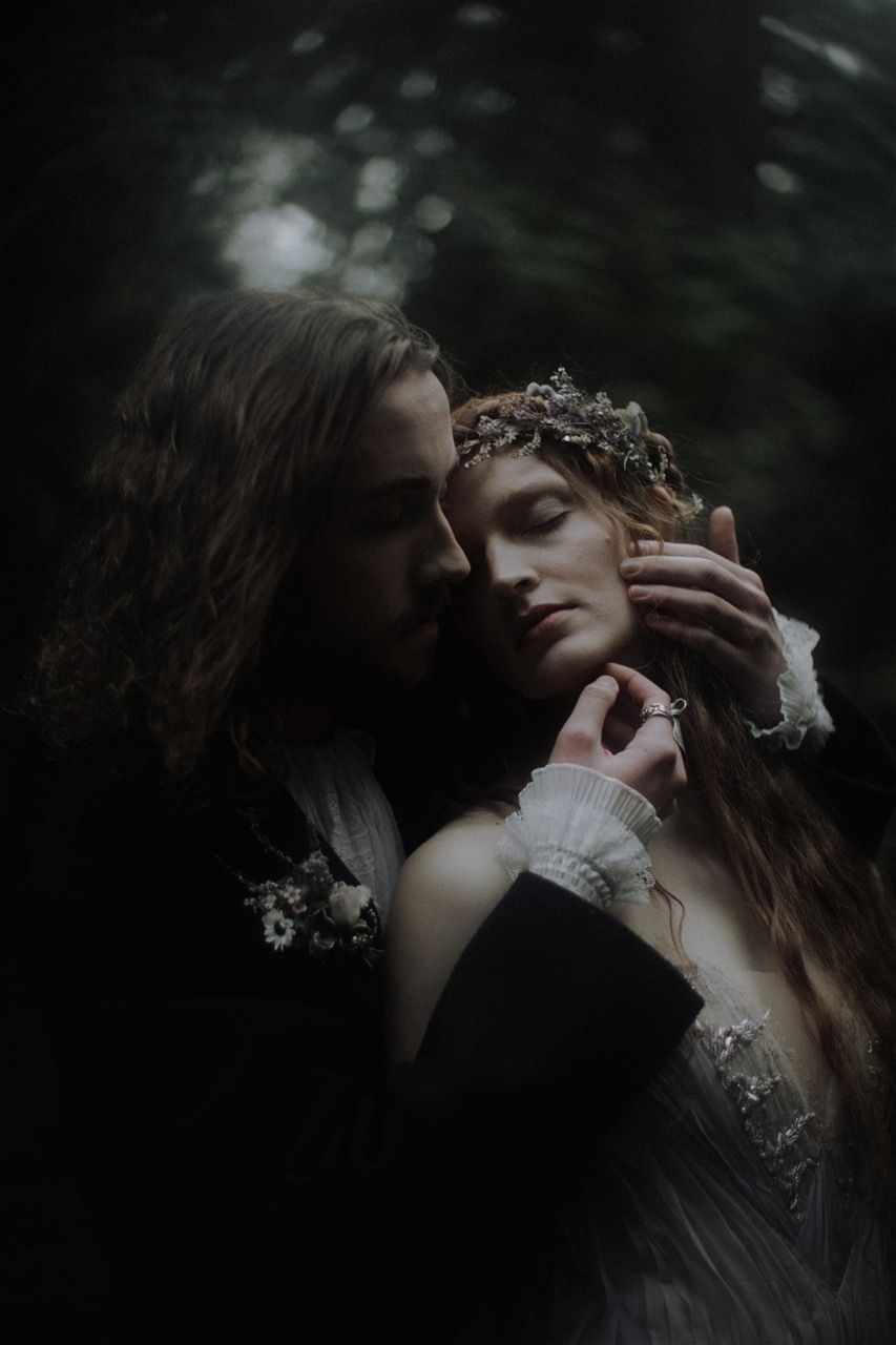 A romantic couple in dark Victorian attire embraces in a moody forest, woman wearing a floral crown with eyes closed.