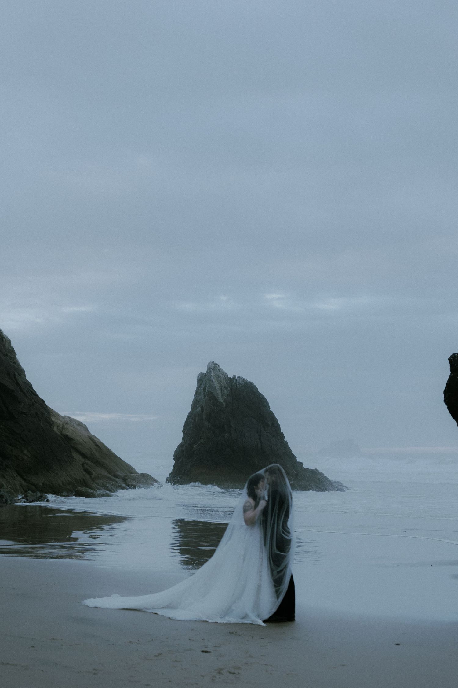 A couple in wedding attire embrace on a misty beach with dramatic rocky cliffs in the background captured by a destination photographer