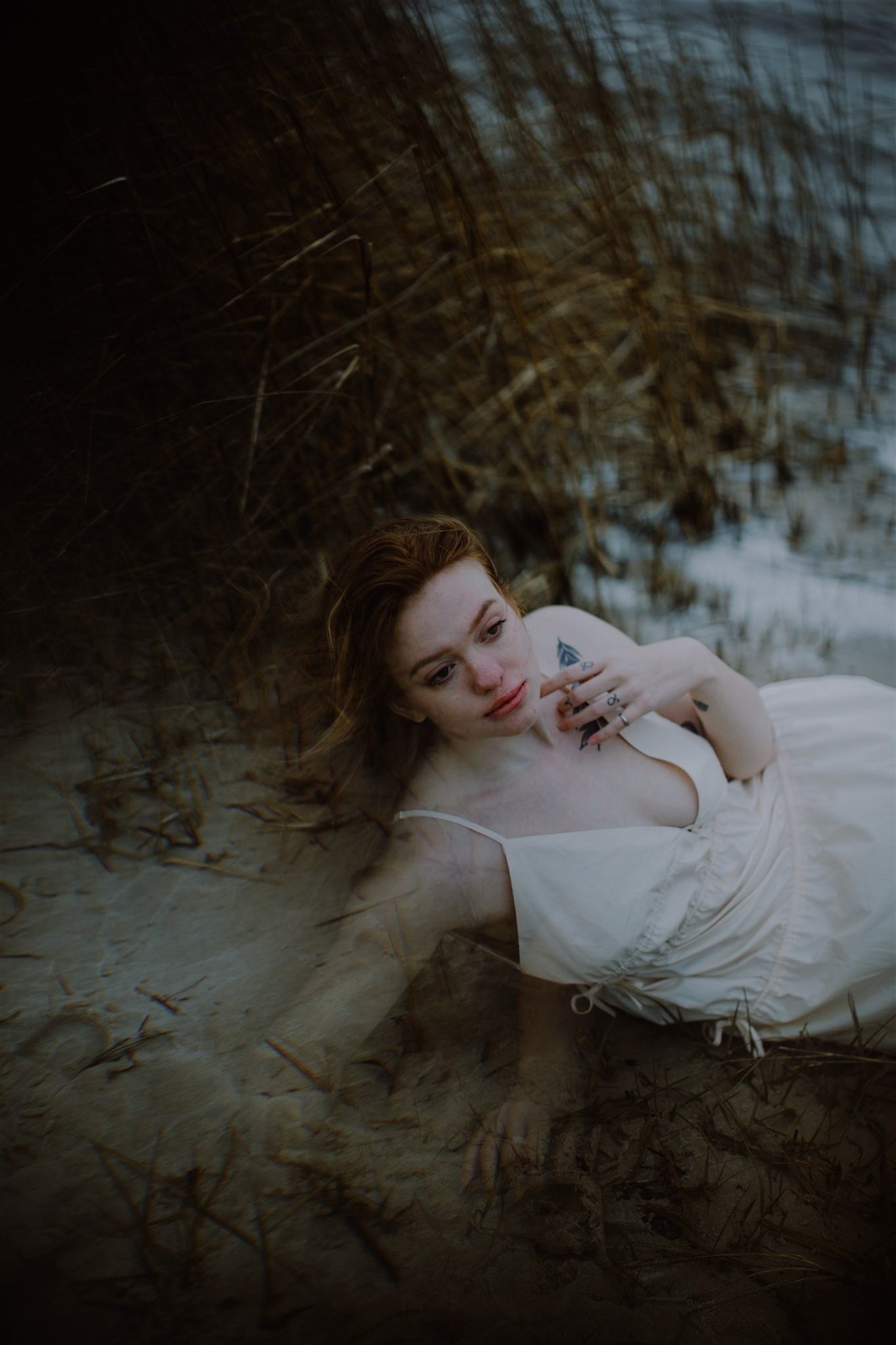 A woman with red hair in a white dress lies on sandy dunes surrounded by tall dry grass in a moody artistic portrait.