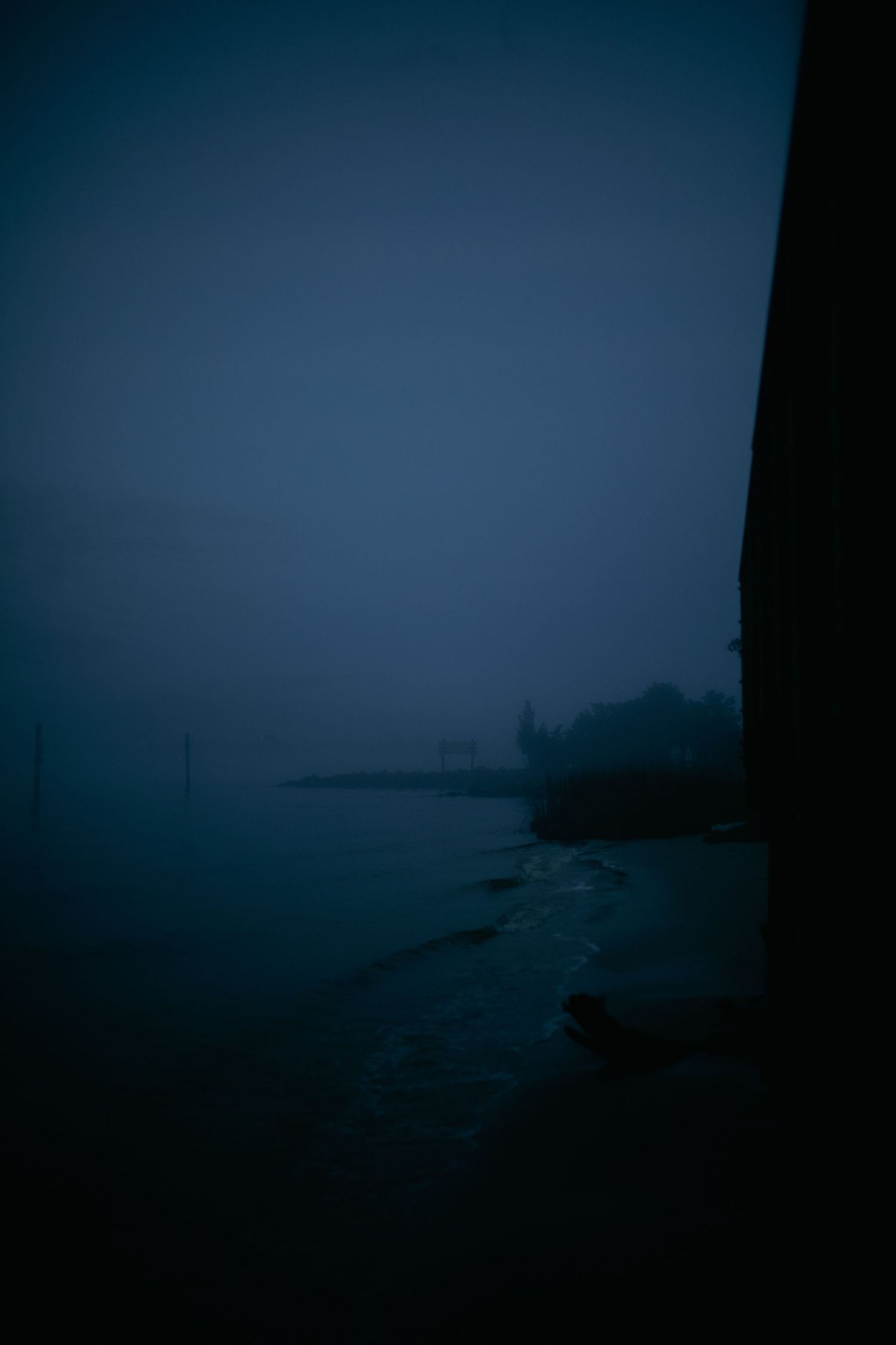 Dark moody coastline shrouded in dense fog at twilight with silhouetted buildings visible in the distance.