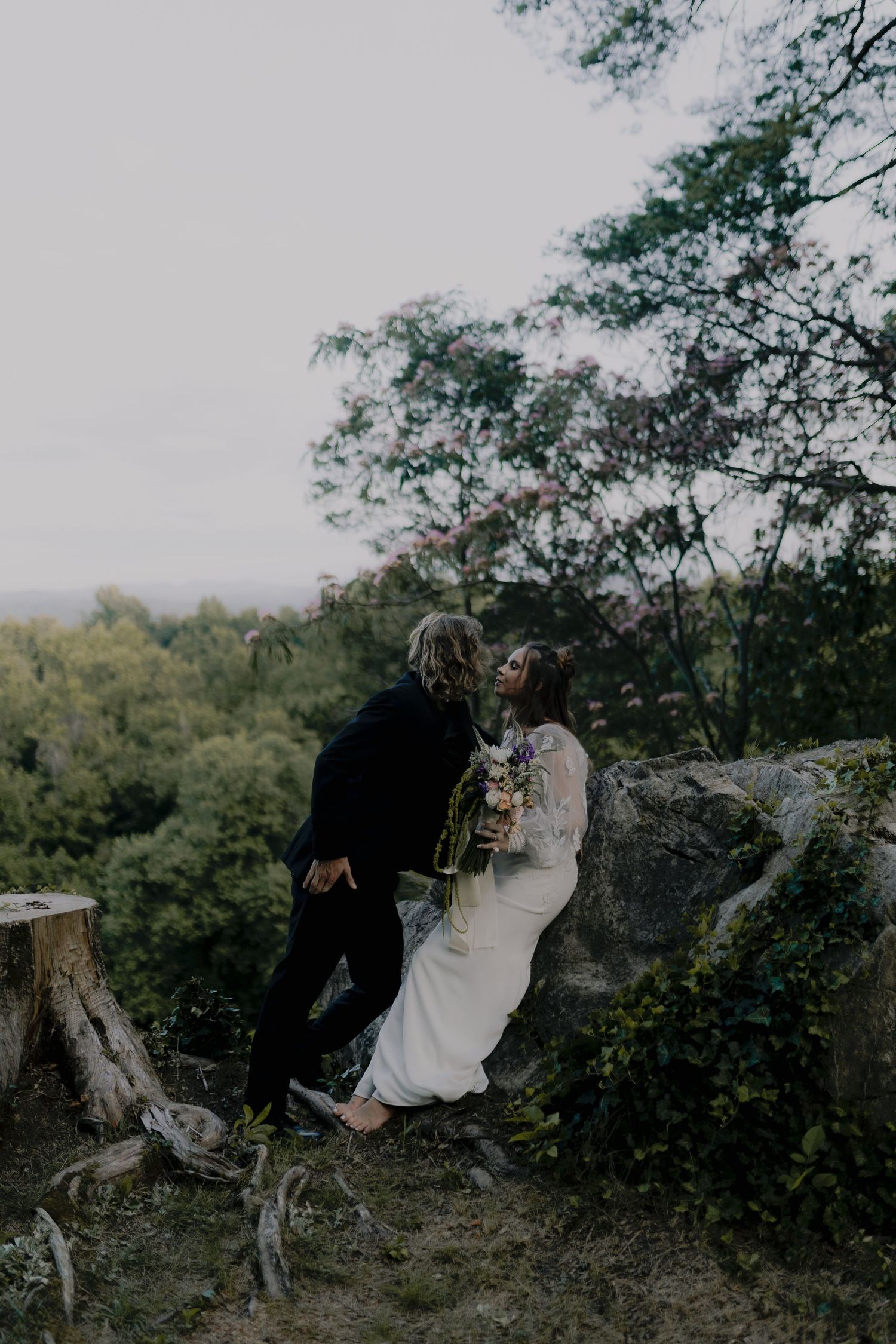 A couple in wedding attire sits together on a rocky cliff edge surrounded by trees during an outdoor photo session.