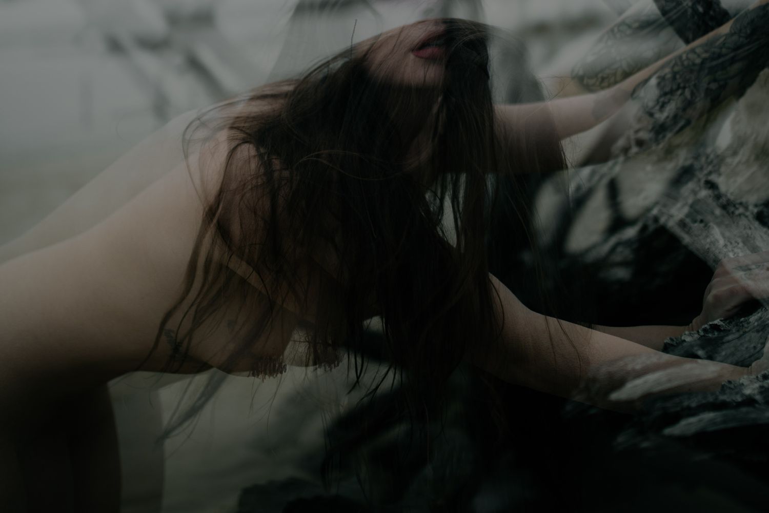 A woman with long dark hair leans forward dramatically among dark rocks in a moody, atmospheric fine art photograph.