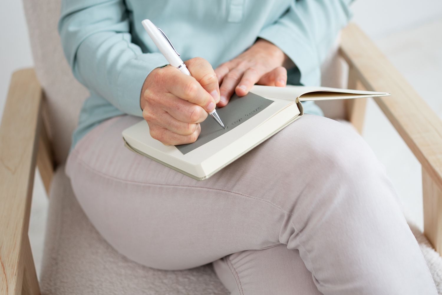 Someone in light blue clothing writing in a notebook while sitting.