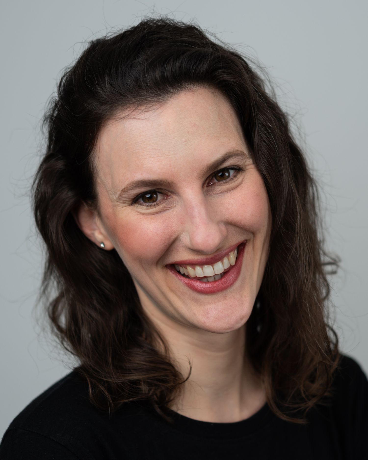 Professional portrait of someone with dark wavy hair wearing a black top against a light background.