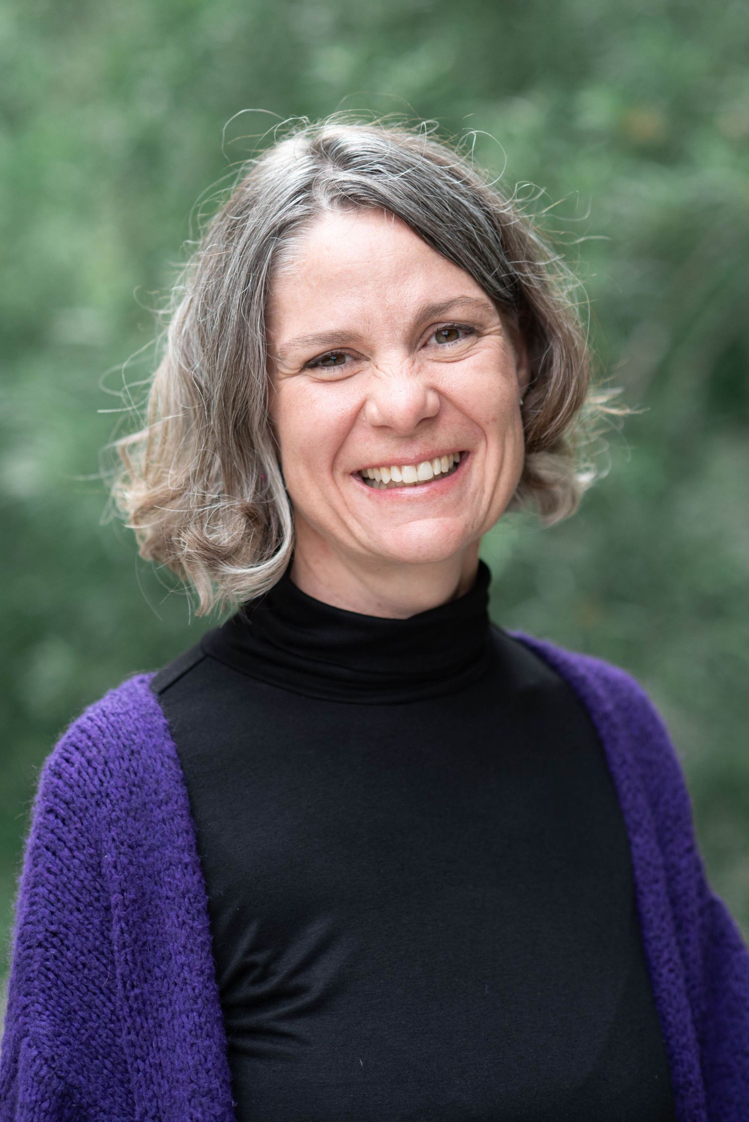 Outdoor portrait of someone wearing a black turtleneck and purple cardigan against a green background.