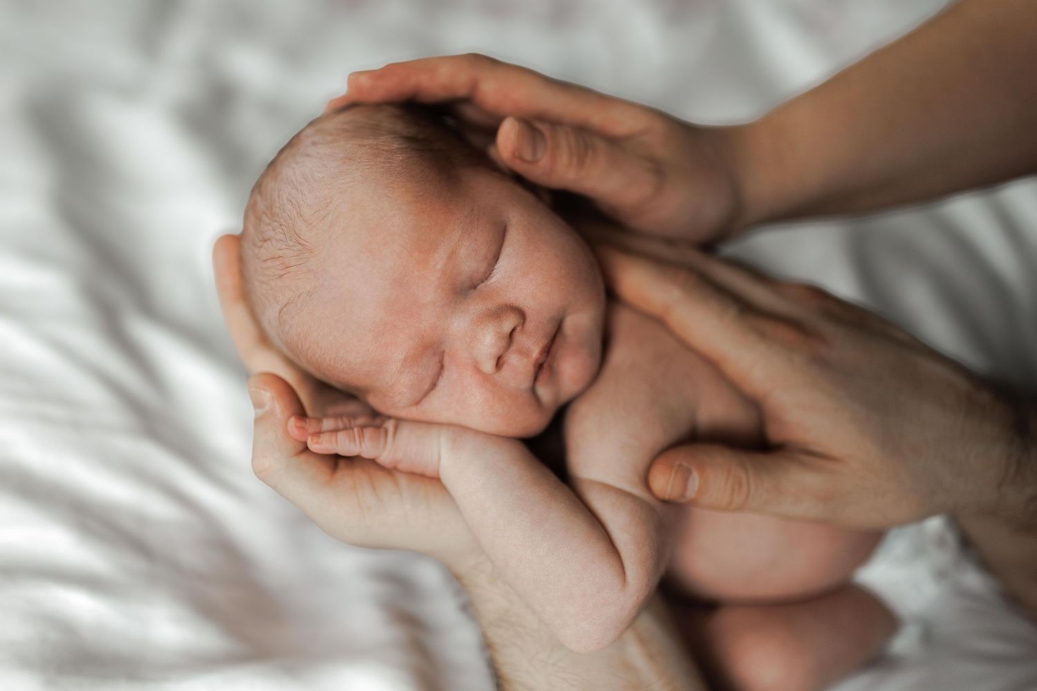 Gentle hands tenderly hold a sleeping newborn baby on soft white bedding.