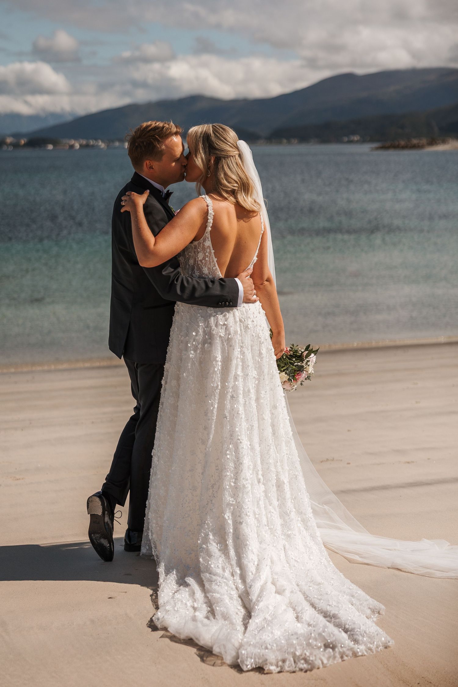 A couple shares a romantic moment on a scenic beach with mountains and water in the background.