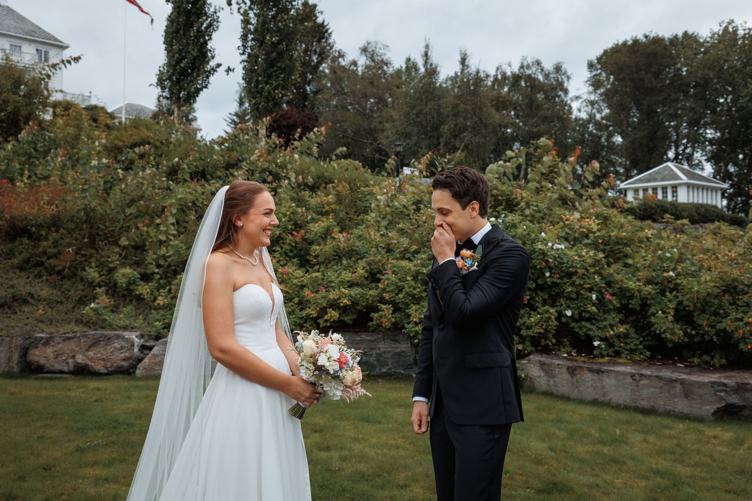 Bride and groom share romantic moments in a garden setting with lush green trees in background.