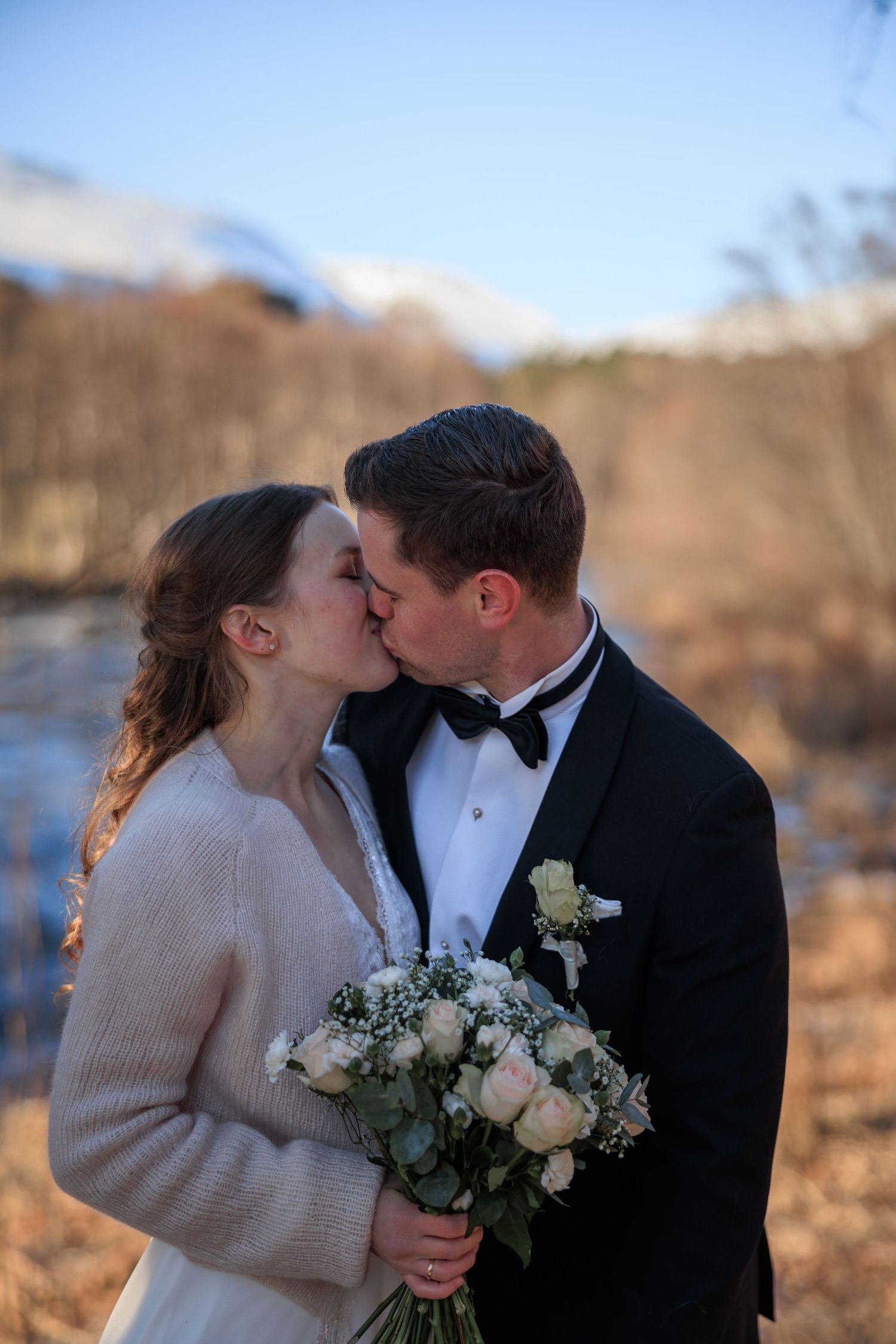 A romantic winter wedding moment with a couple sharing a kiss as the bride holds a white floral bouquet.