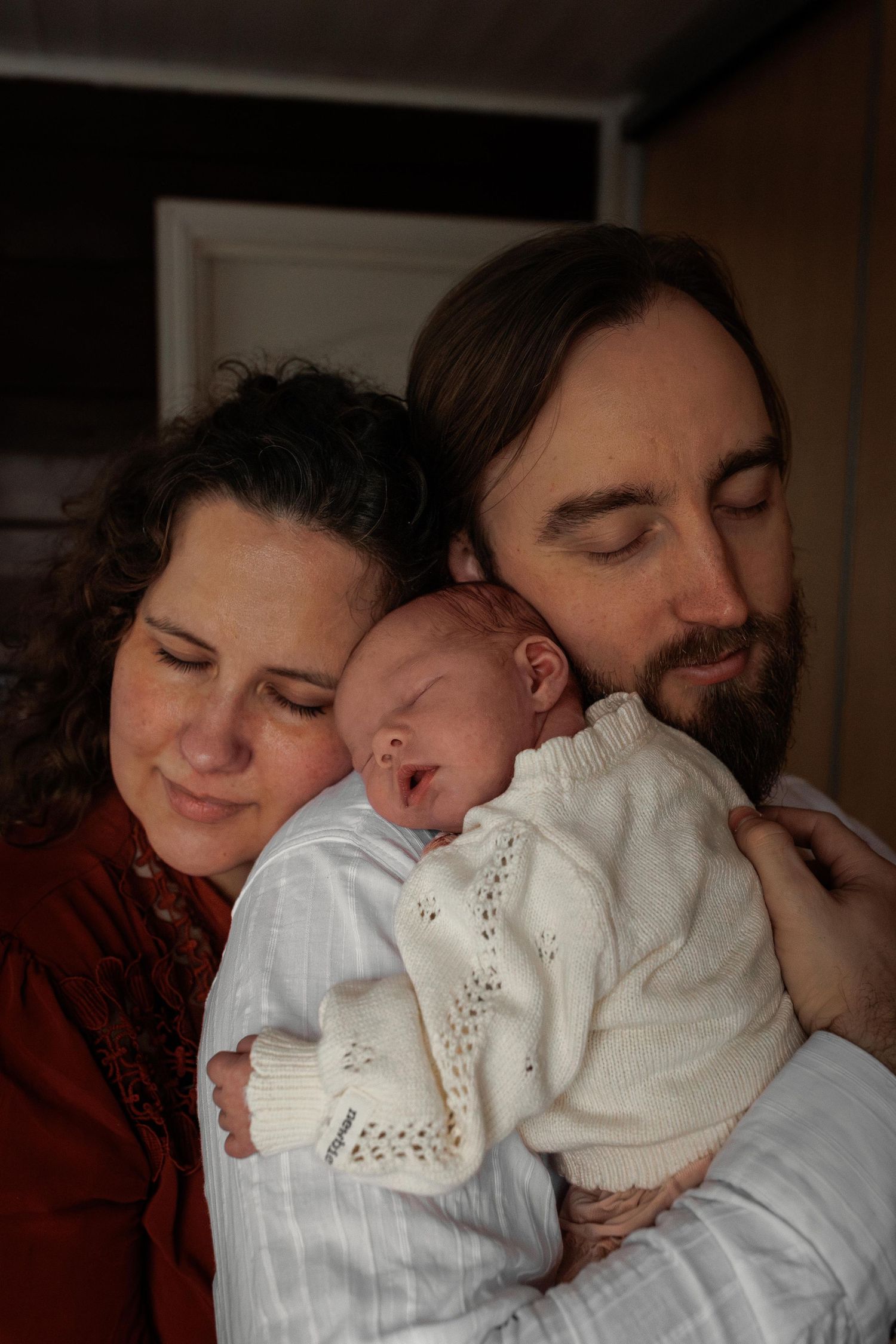 A sleeping newborn baby in white clothing is held lovingly by a couple in a warm, intimate indoor setting.
